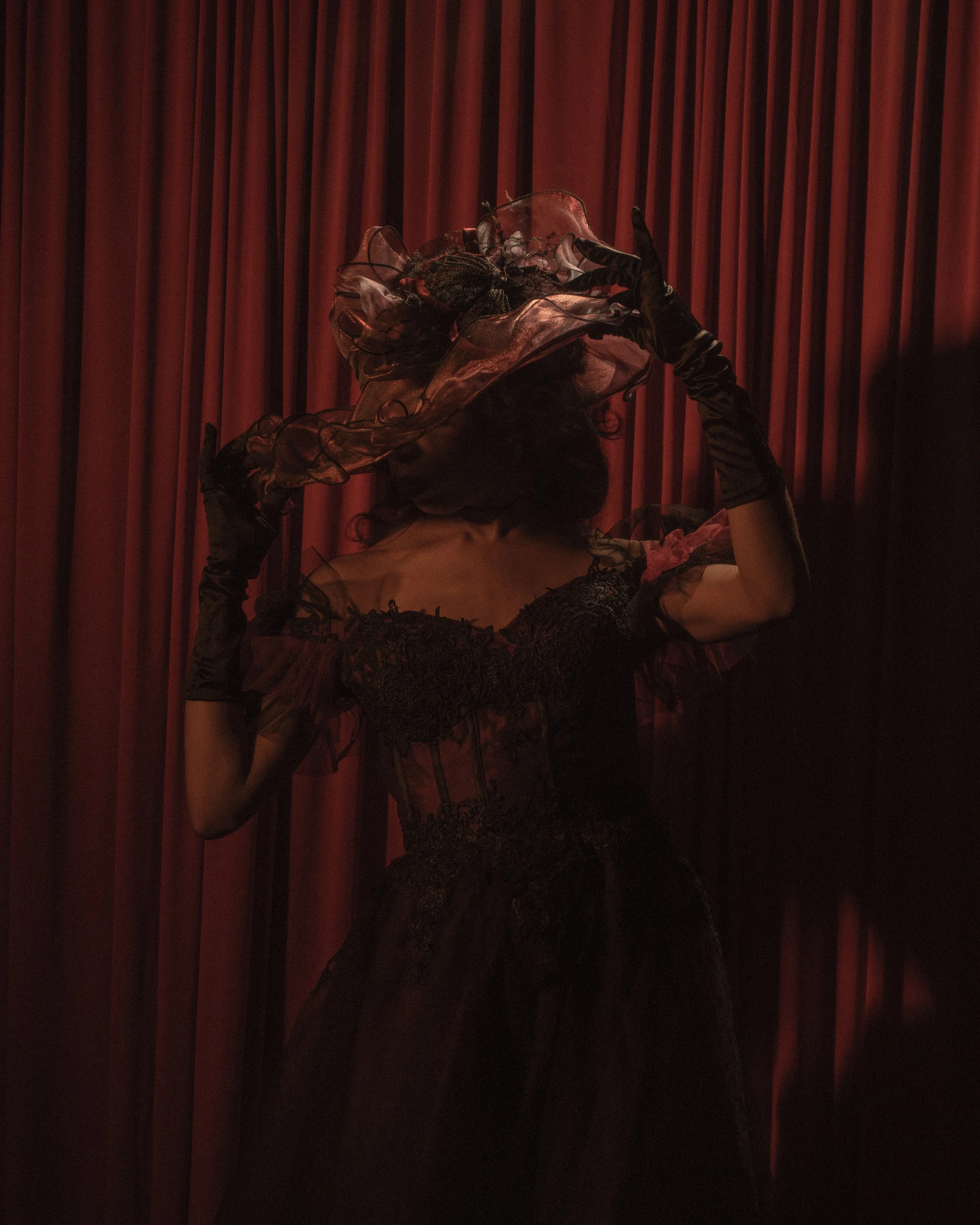 A person dressed in black with long gloves and a fancy hat is sitting in front of red velvet curtains.