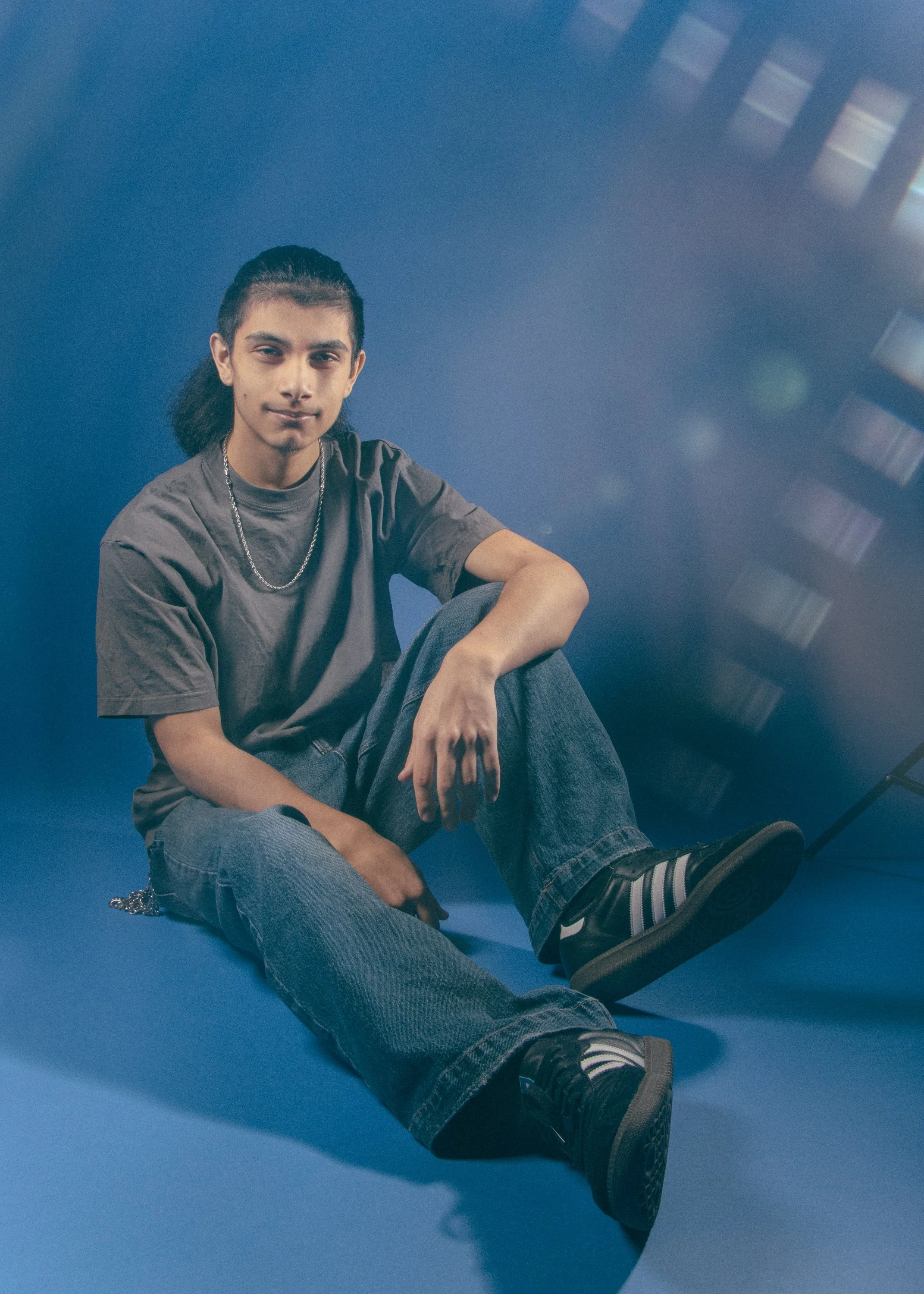 A young man with long dark hair tied back, wearing a gray t-shirt, jeans, and sneakers, sitting on a blue floor with one knee up, looking at the camera, with a blue background and a soft light effect.