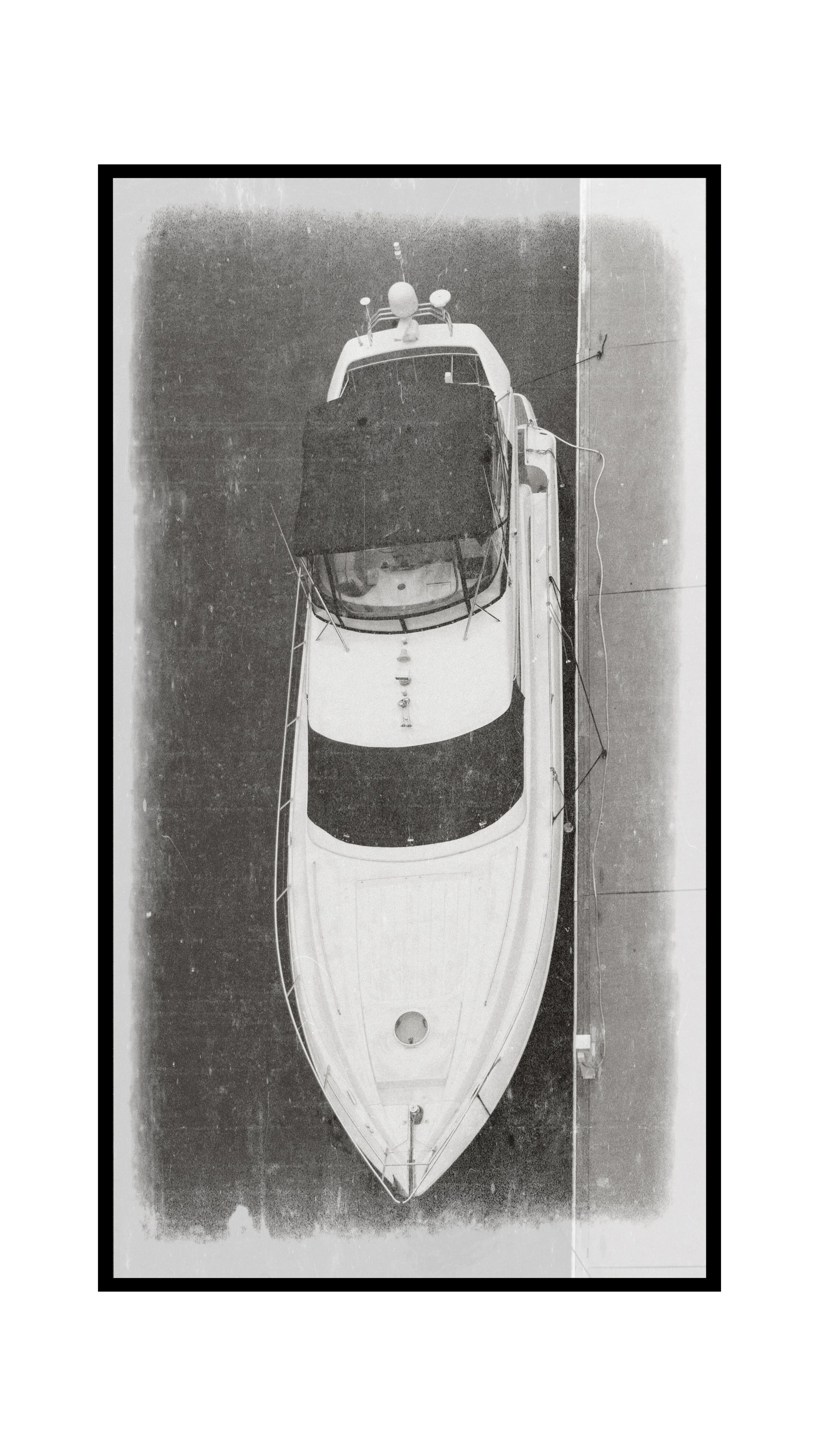 Black and white photograph of a boat viewed from above, positioned on a dock with visible ropes securing it.