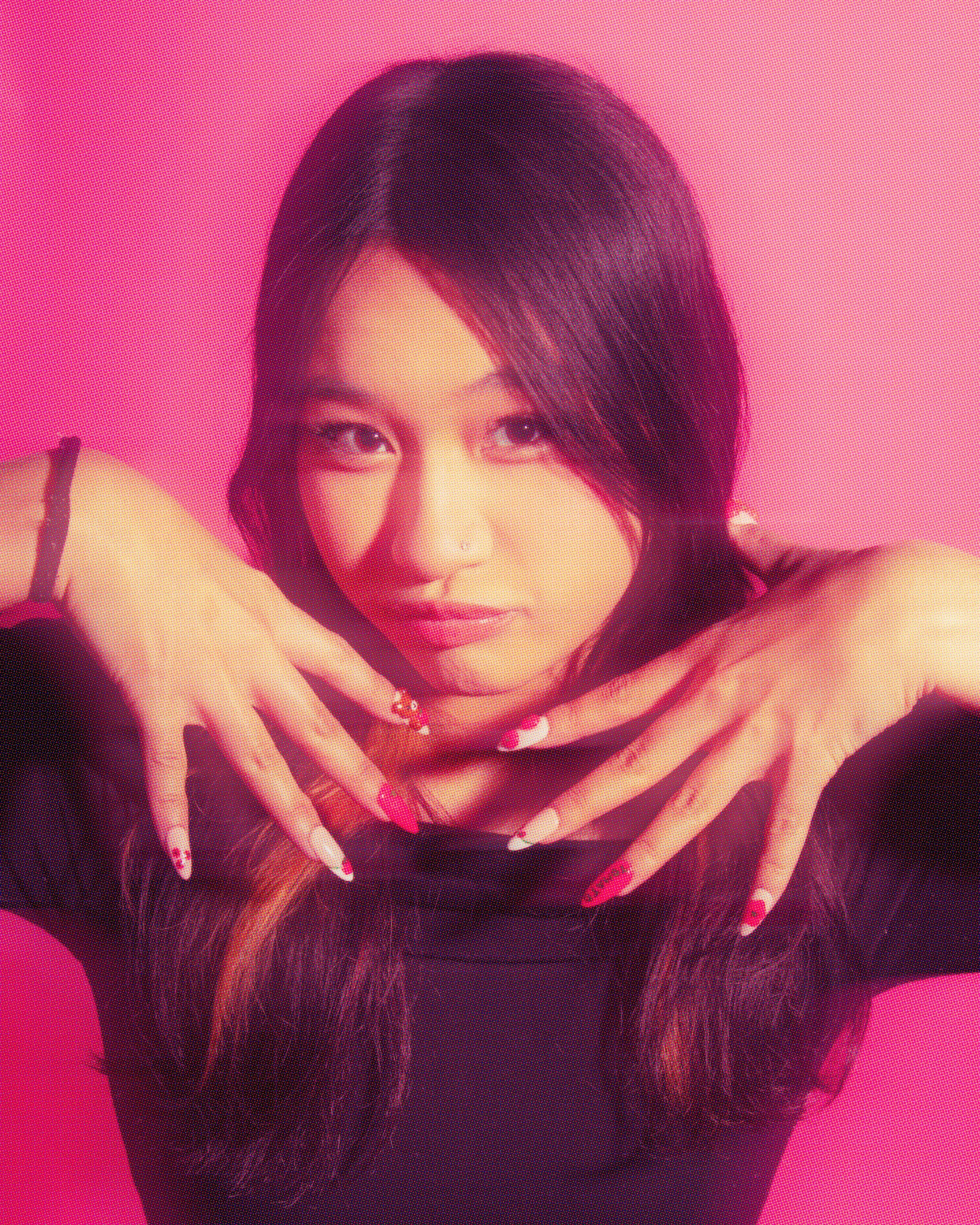 A young woman with long dark hair, wearing a black top, posing with her hands near her face against a pink background.