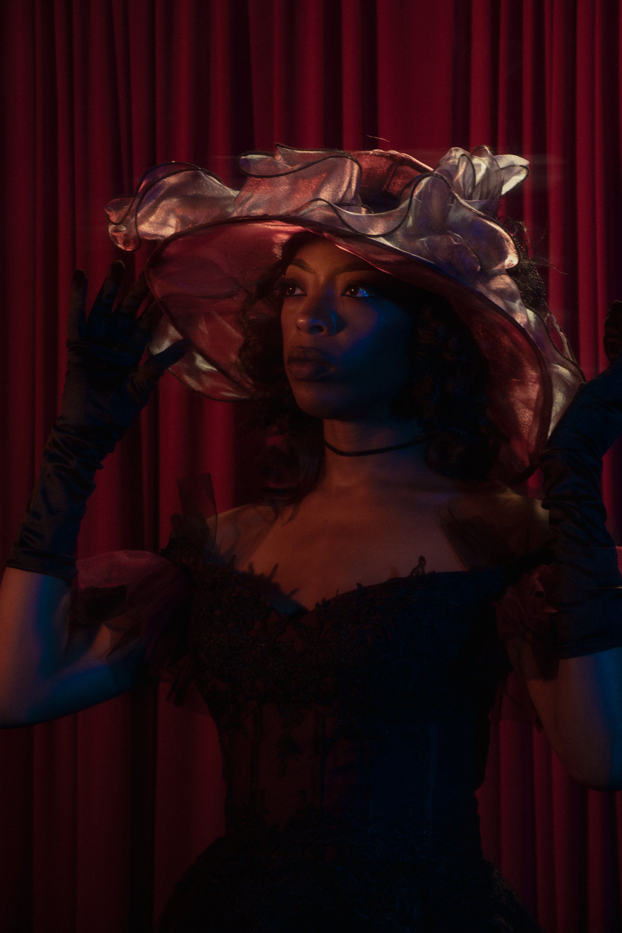 A woman dressed in a black gown and long black gloves, wearing a large, ruffled, semi-transparent hat, stands in front of red curtains, with dramatic lighting highlighting her face.