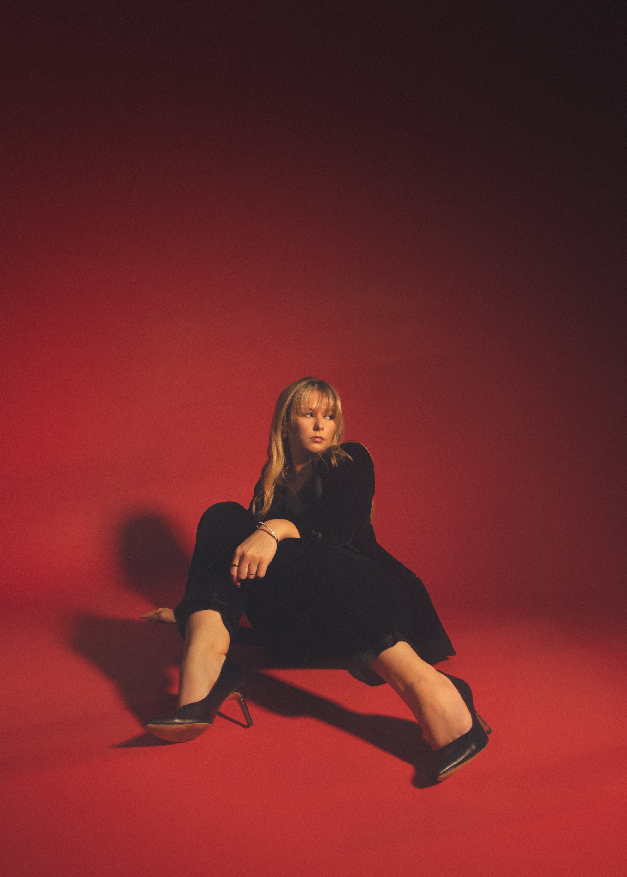 A woman with long blonde hair, wearing a black outfit and high-heeled shoes, is sitting on the floor against a red background. She has a serious expression and is resting her chin on her hand.