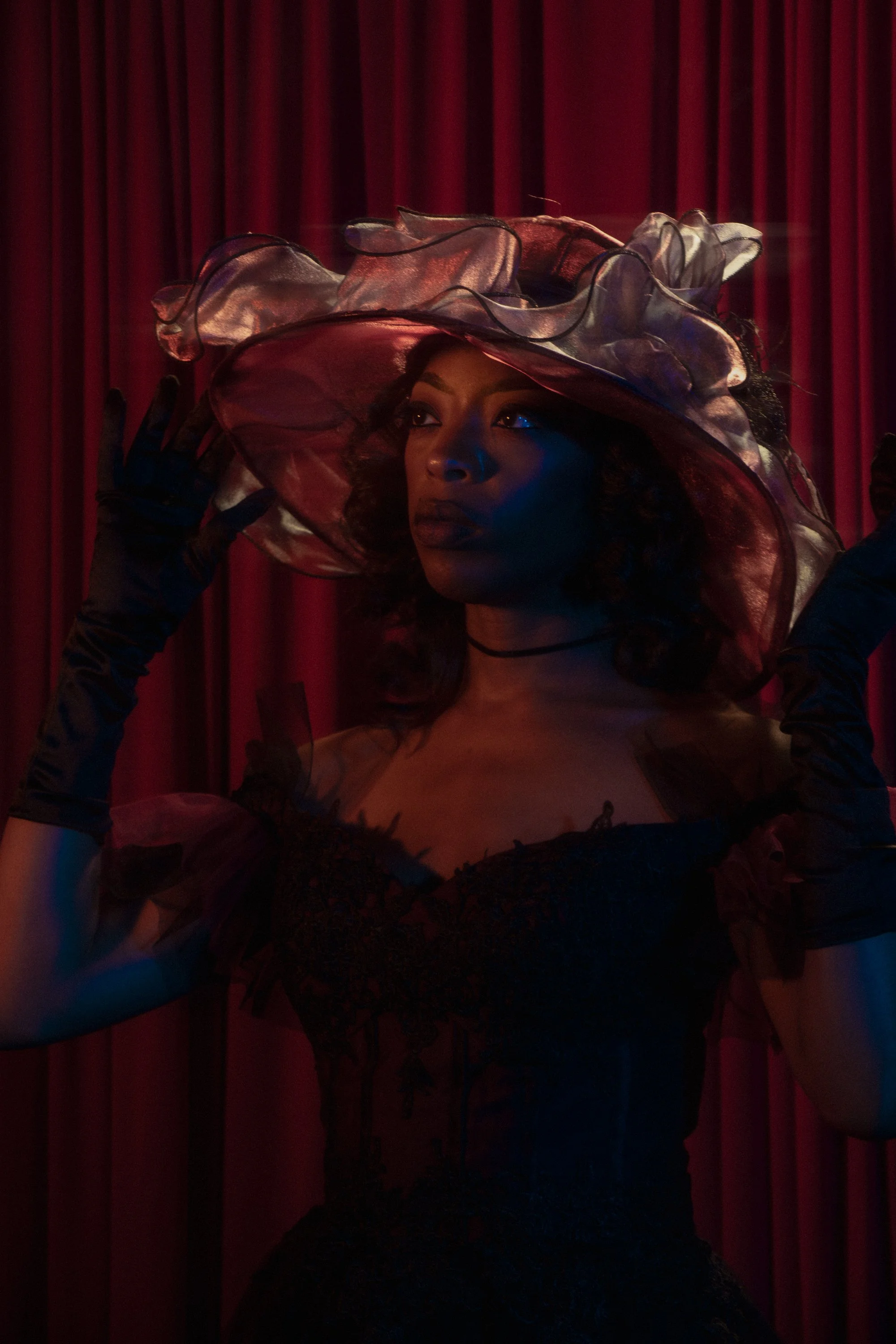 A woman with dark skin and curly hair wearing a large, colorful, ruffled hat with sheer fabric and black accents, posing against a red velvet curtain backdrop.