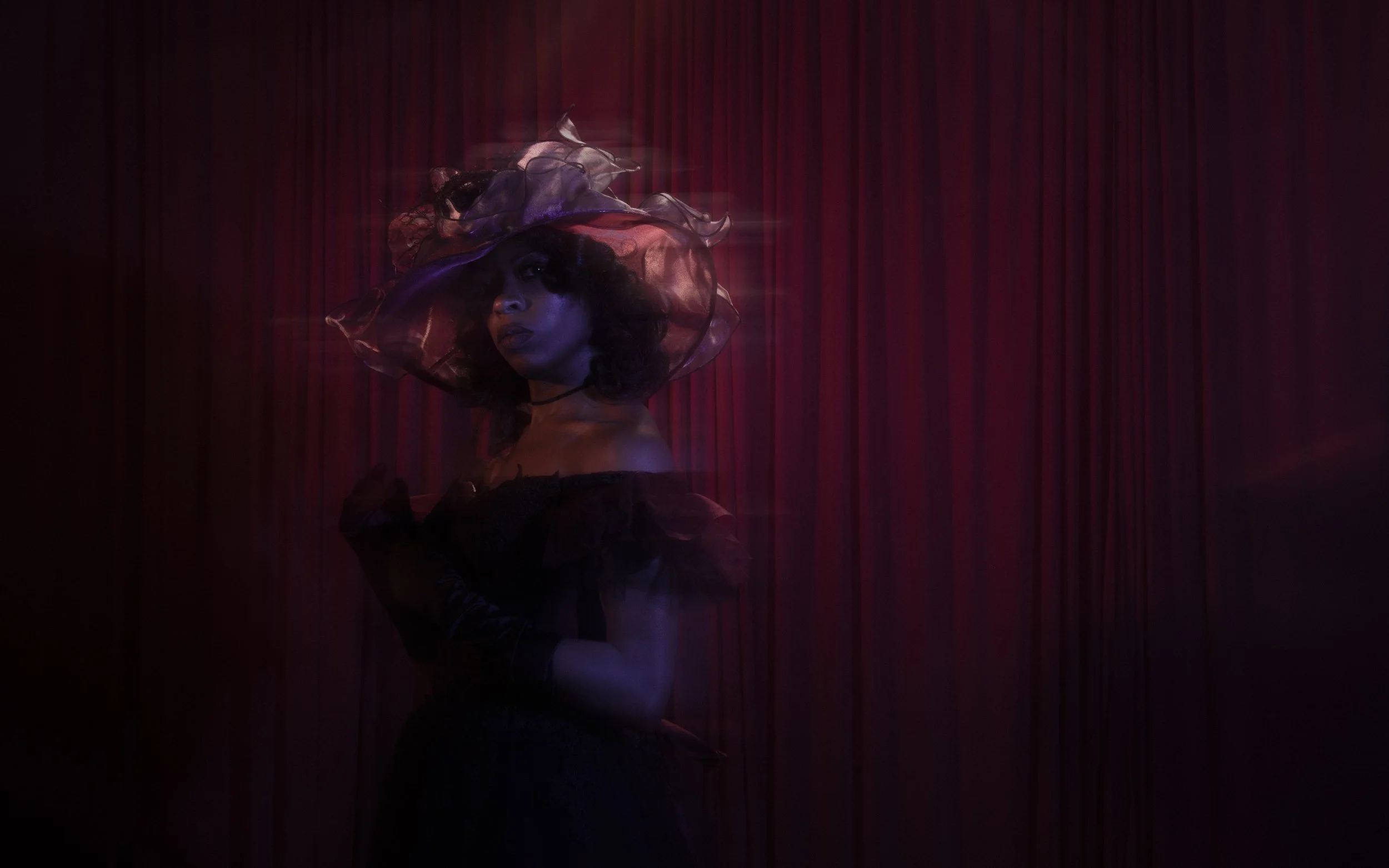 A person with curly hair wearing a large, colorful, sheer hat against a dark red curtain background.