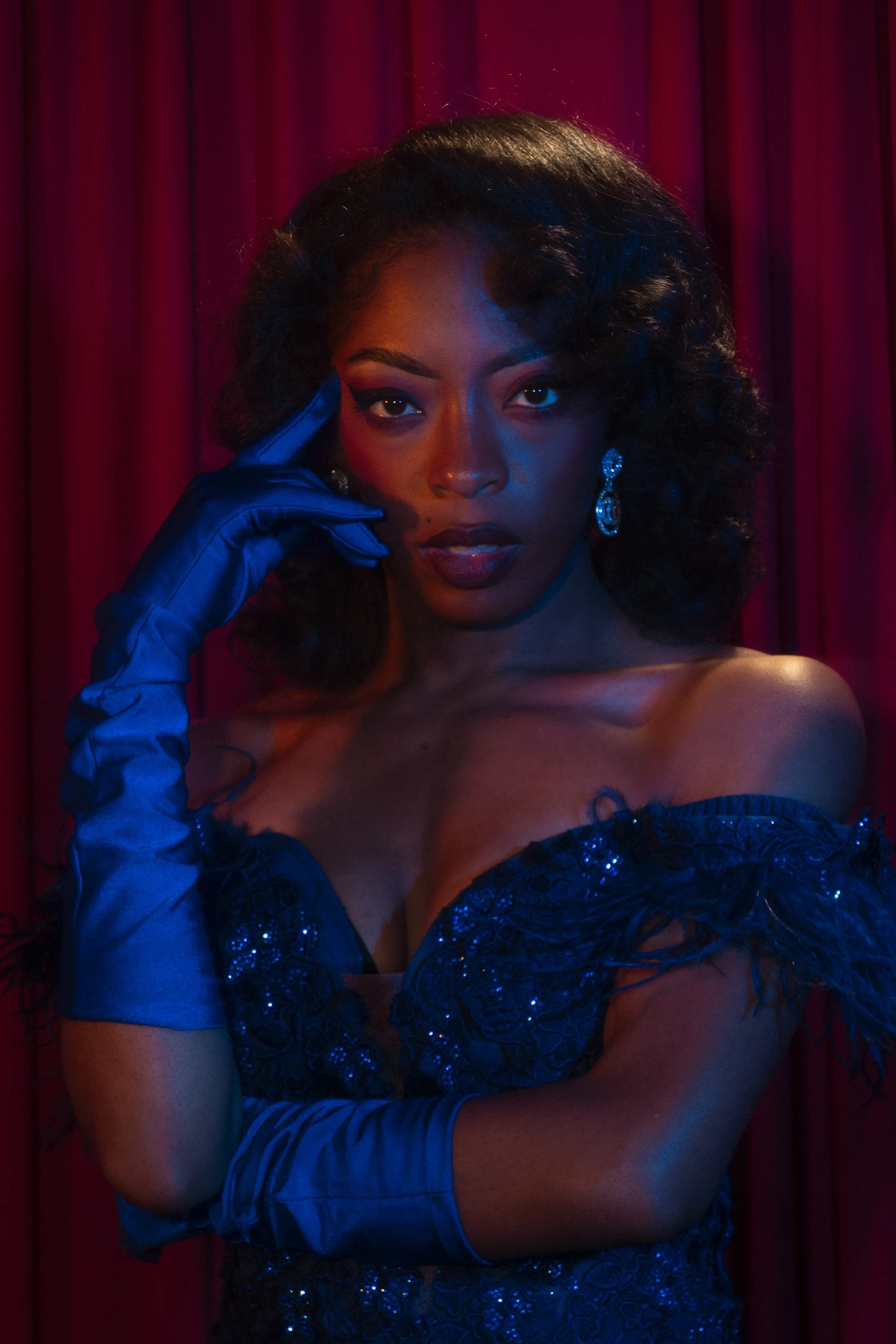 A woman in a strapless, dark blue, embroidered gown with sequins, wearing long blue gloves, and large sparkling earrings. She poses with one hand touching her temple against a backdrop of red curtains, with dramatic lighting emphasizing her makeup an