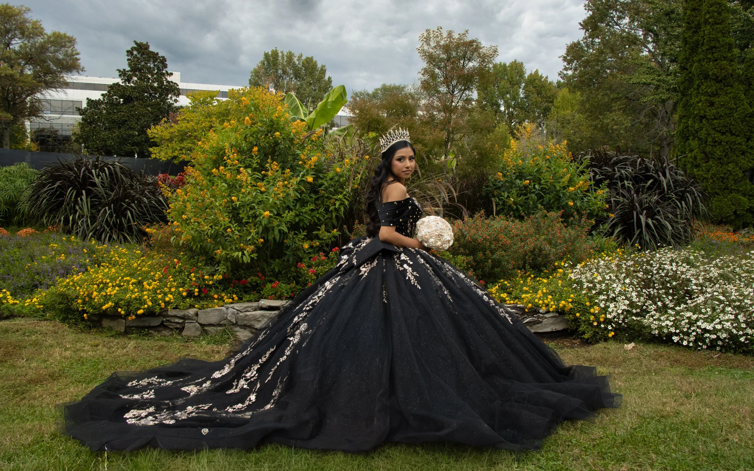 A woman in an elegant black gown with white floral patterns, wearing a silver tiara, standing in a garden with yellow and white flowers, holding a bouquet of white roses, overcast sky in the background.