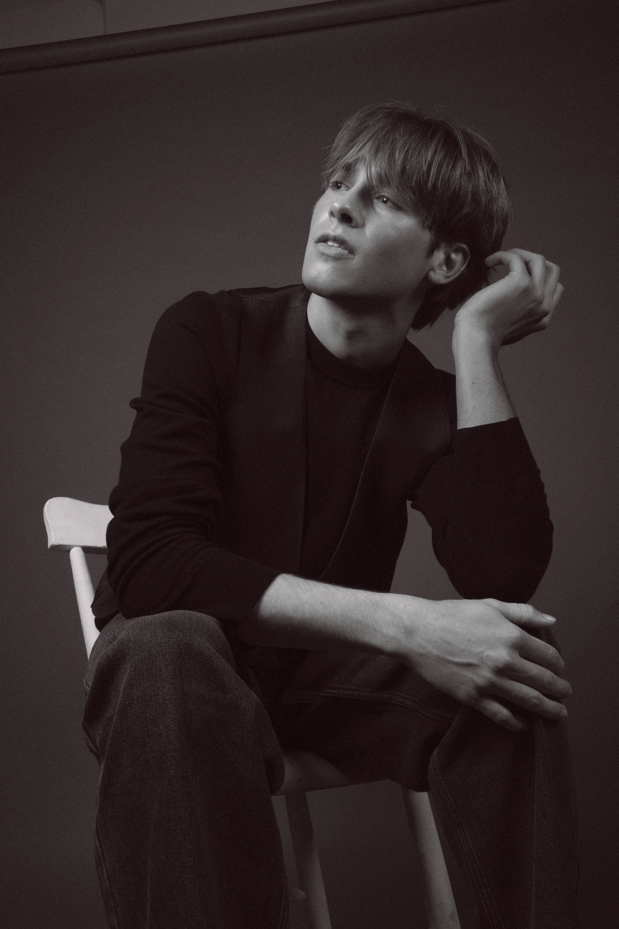 A black and white photo of a young man with medium-length hair, sitting on a white chair, resting his head on his left hand, looking pensively to the side.
