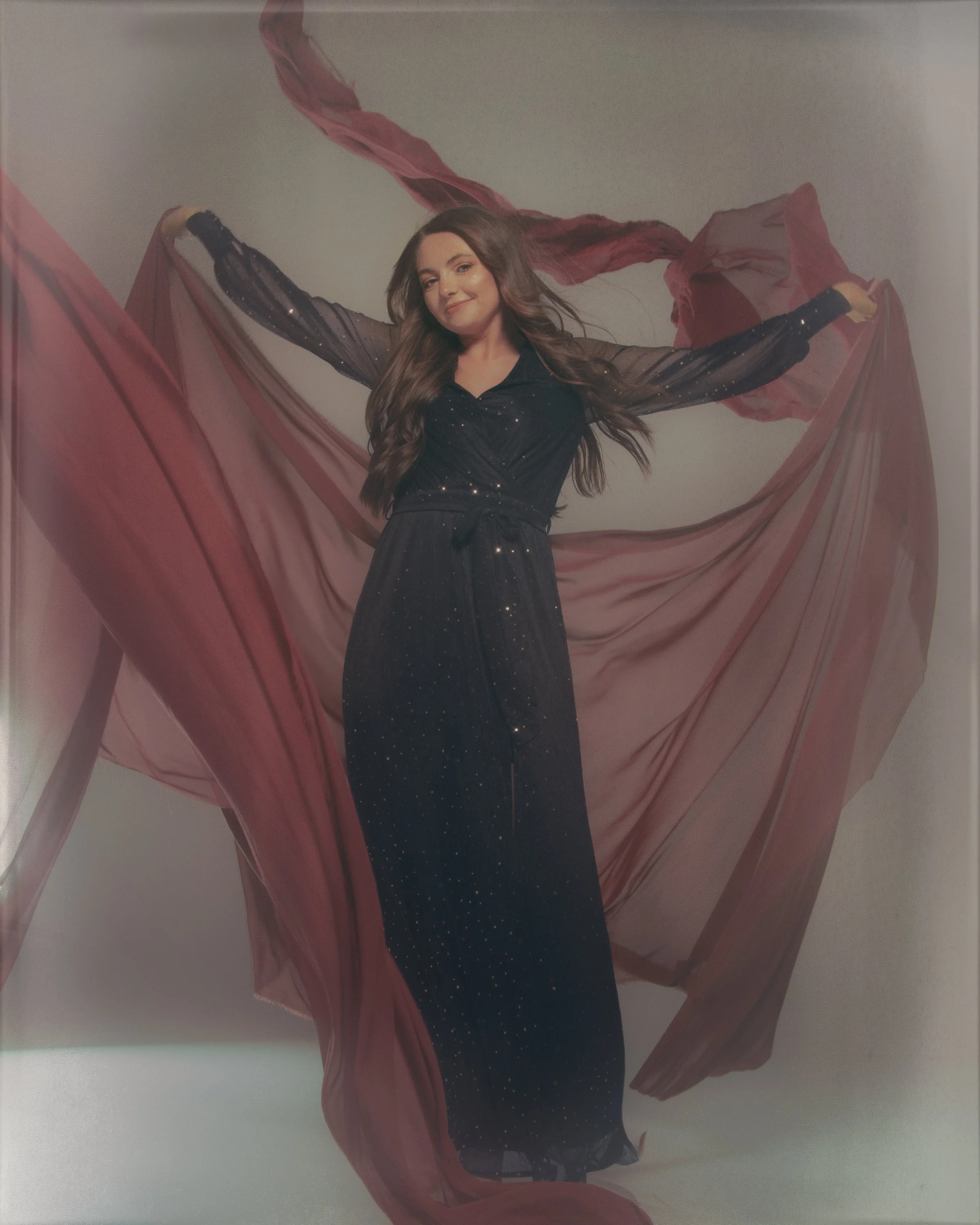 A young woman with long brown hair wearing a black dress with sheer, polka-dotted sleeves and holding large, flowing pink fabric in both hands, standing against a plain, light-colored background.