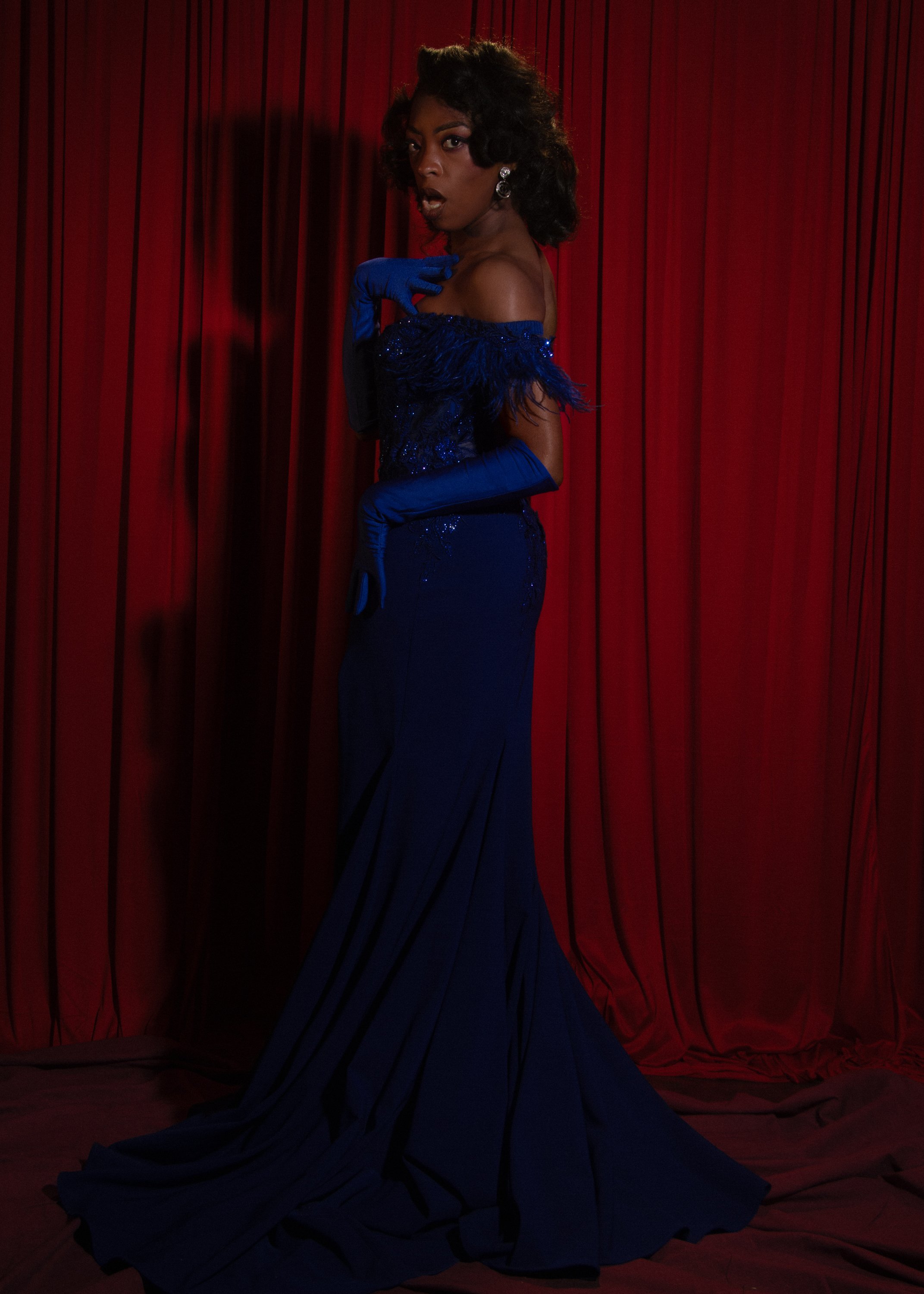 A woman in a blue gown and matching gloves posing in front of red curtains.