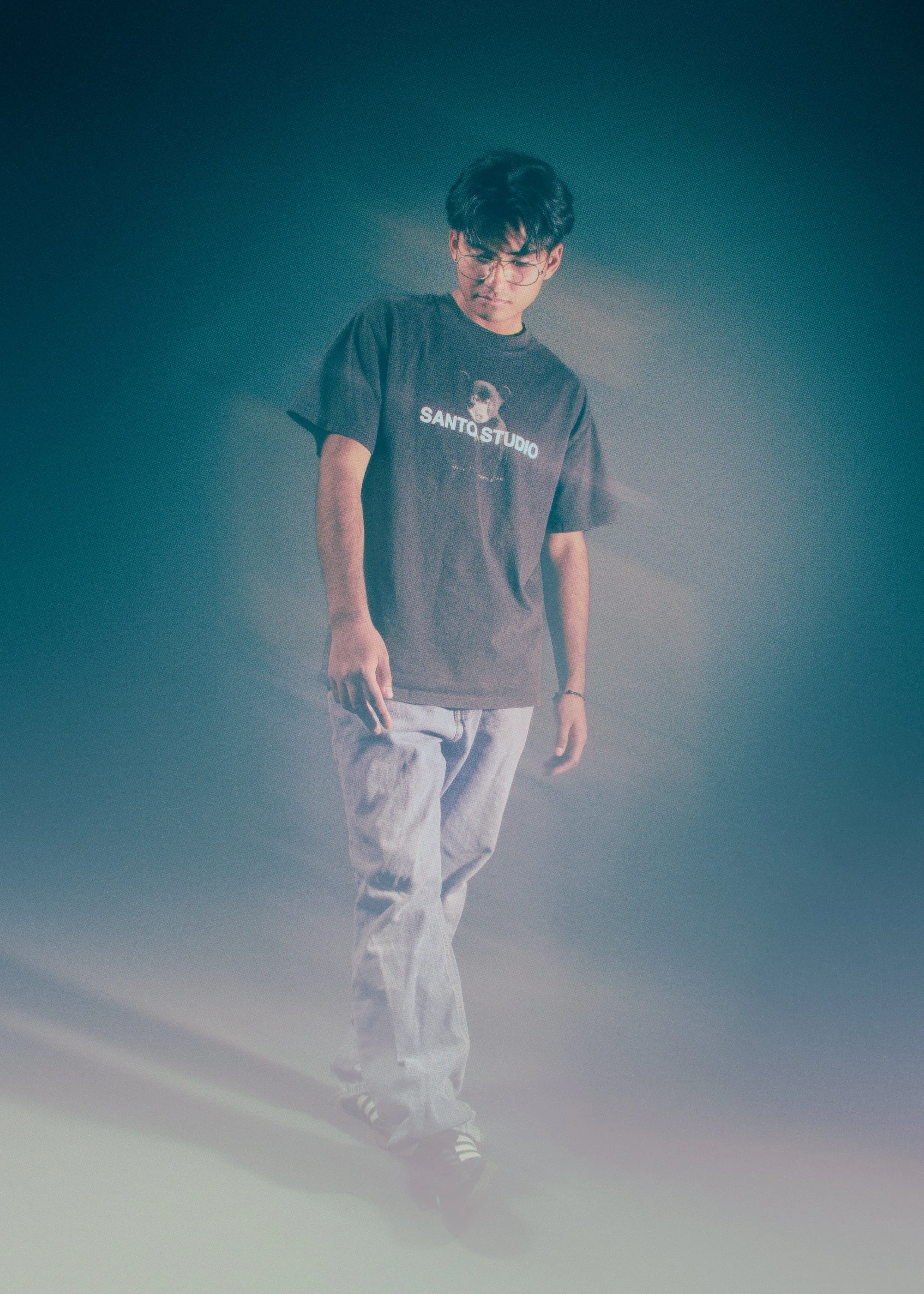 A young man with curly dark hair and glasses looking downward, wearing a black T-shirt with a graphic and the words 'SANTO STUDIO', light-colored pants, and black and white sneakers, standing on a foggy or smoky stage with dramatic lighting.