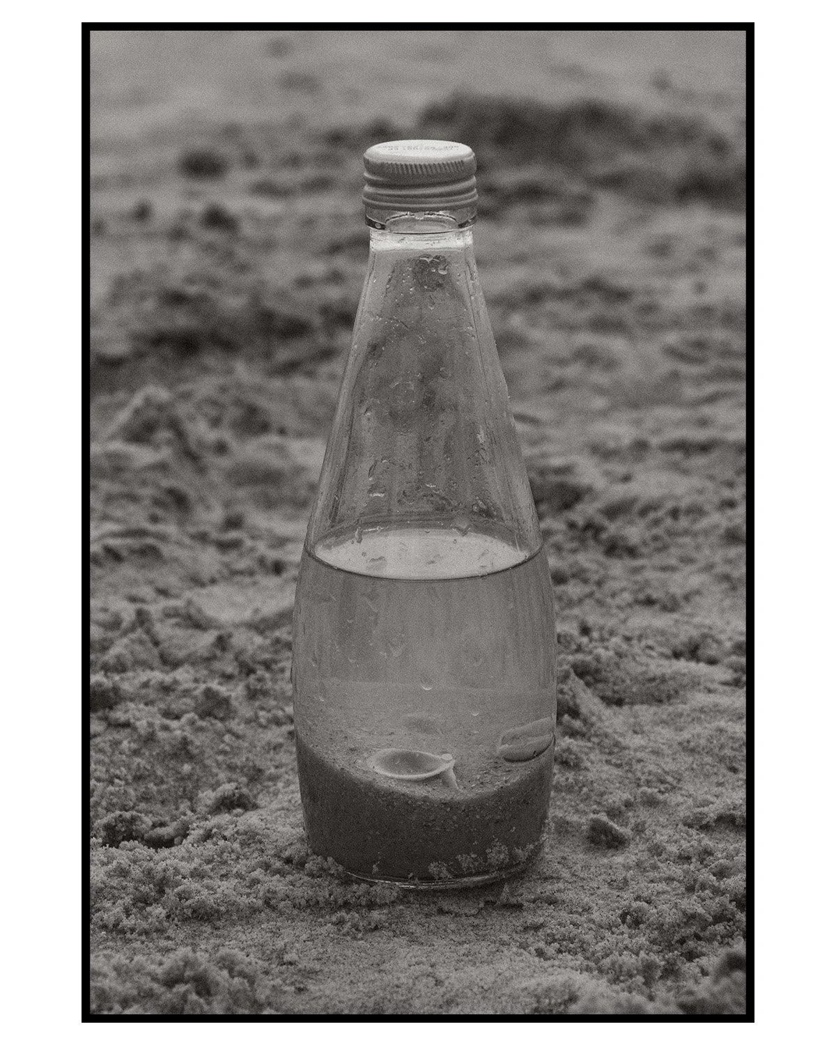 A glass bottle filled with water, placed on sandy ground, with a mountain or hill in the background.