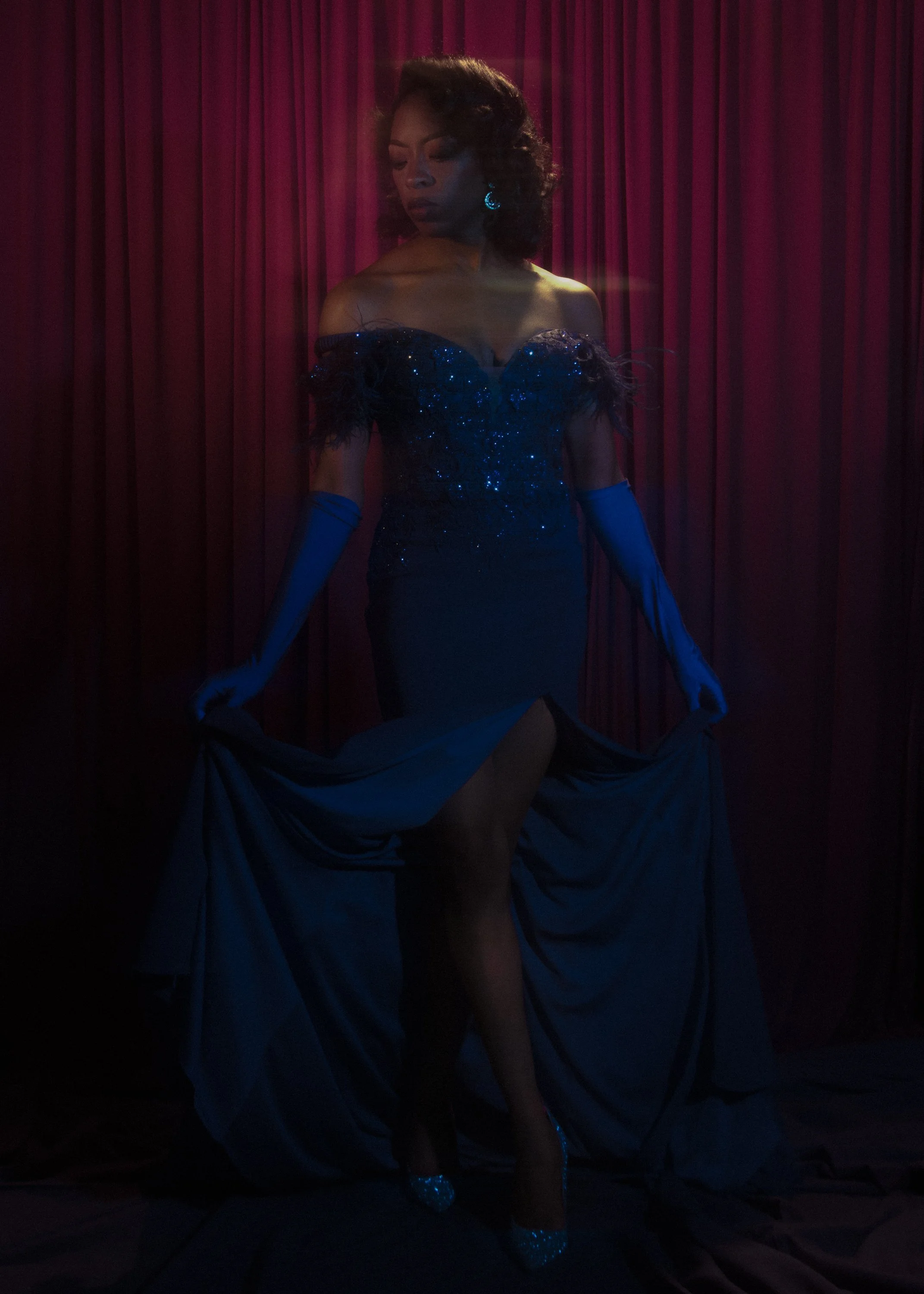 A woman in a glamorous, sparkling, off-shoulder dress with feather accents and long gloves, standing against a backdrop of red velvet curtains at a performance or event.