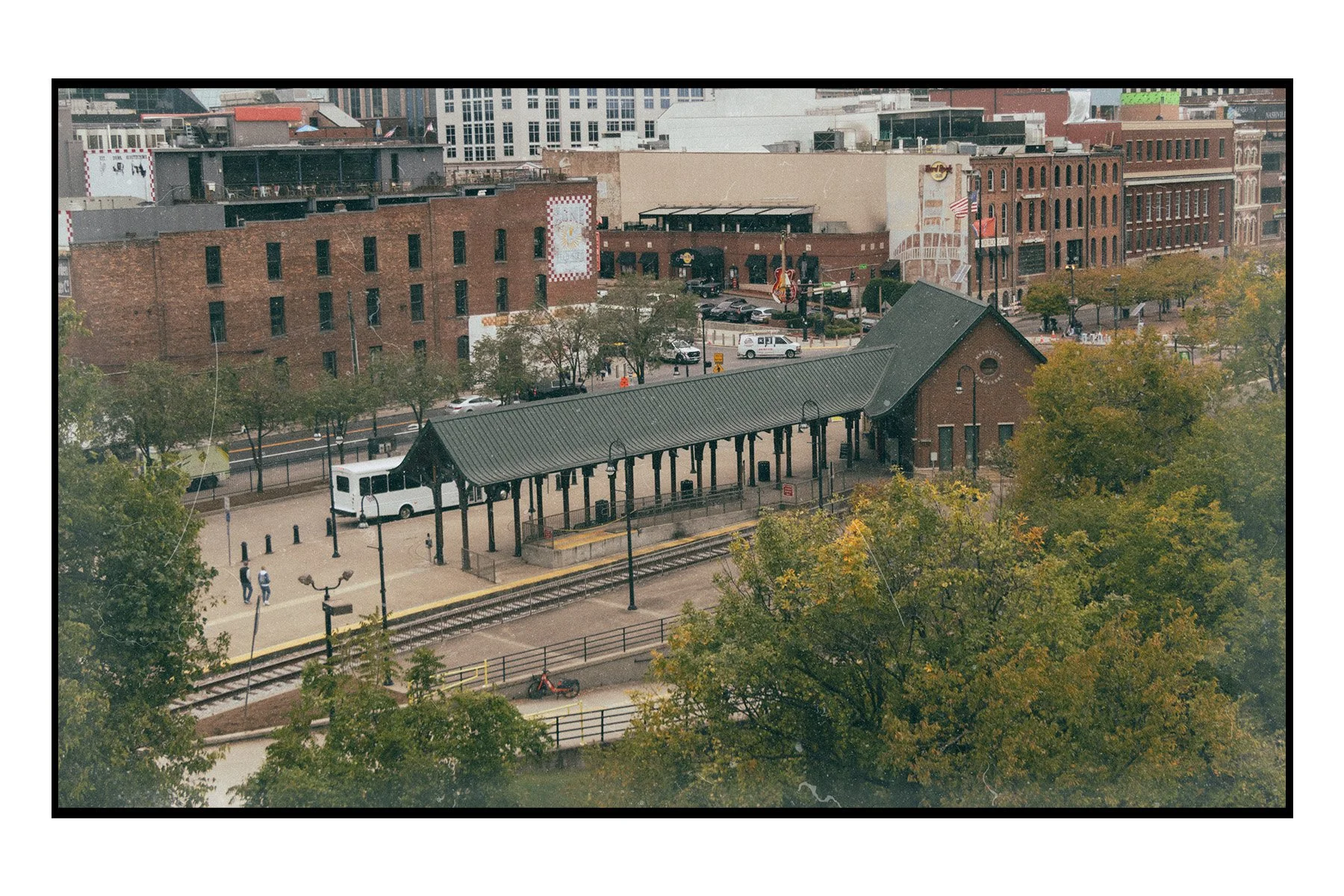 View of an outdoor train station platform with tracks, a small brick building, and a covered waiting area with benches, surrounded by trees with autumn leaves and city buildings in the background.