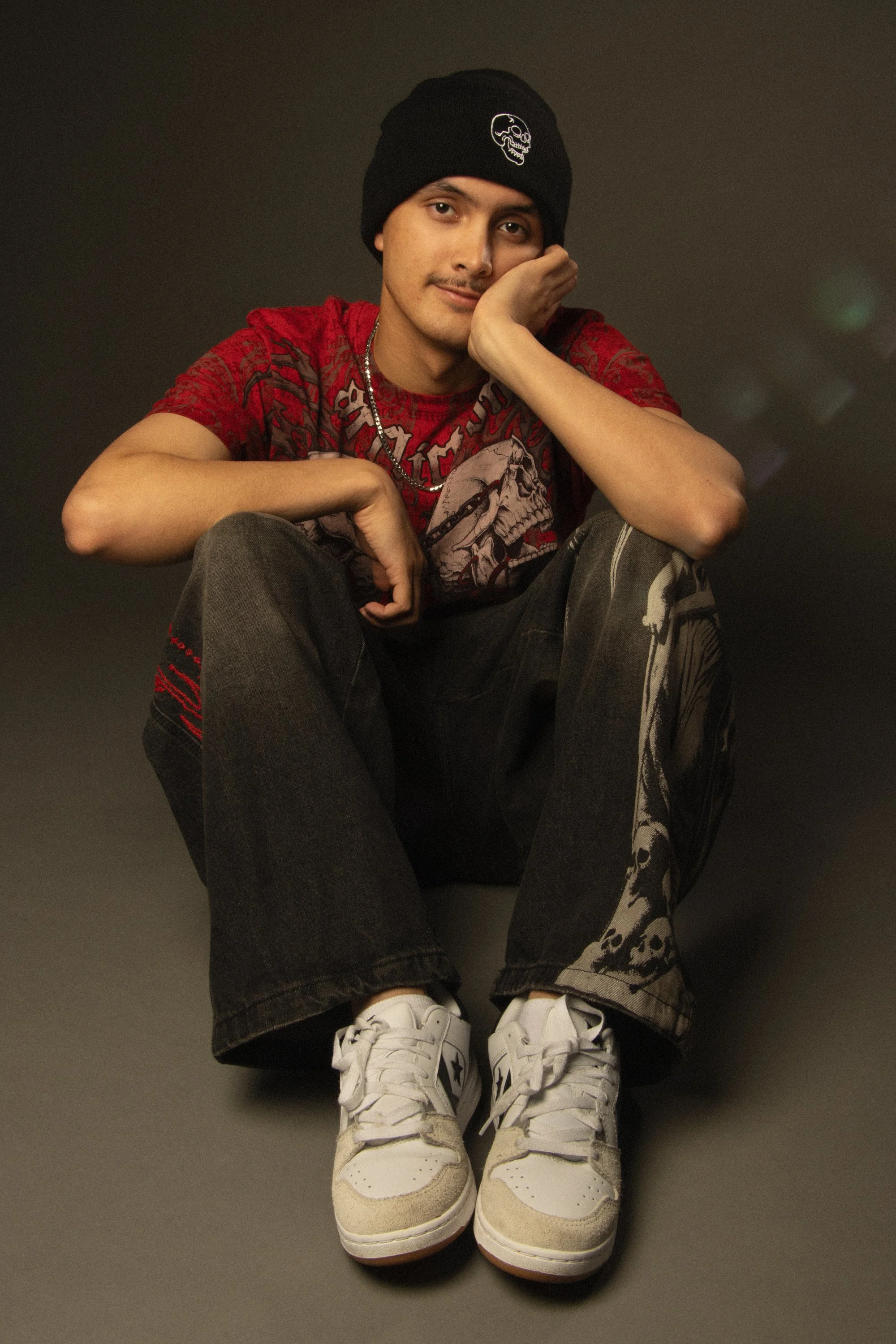A young man sitting on the floor, wearing a black beanie with a skull logo, a red graphic t-shirt, black jeans, and white sneakers, against a dark background.