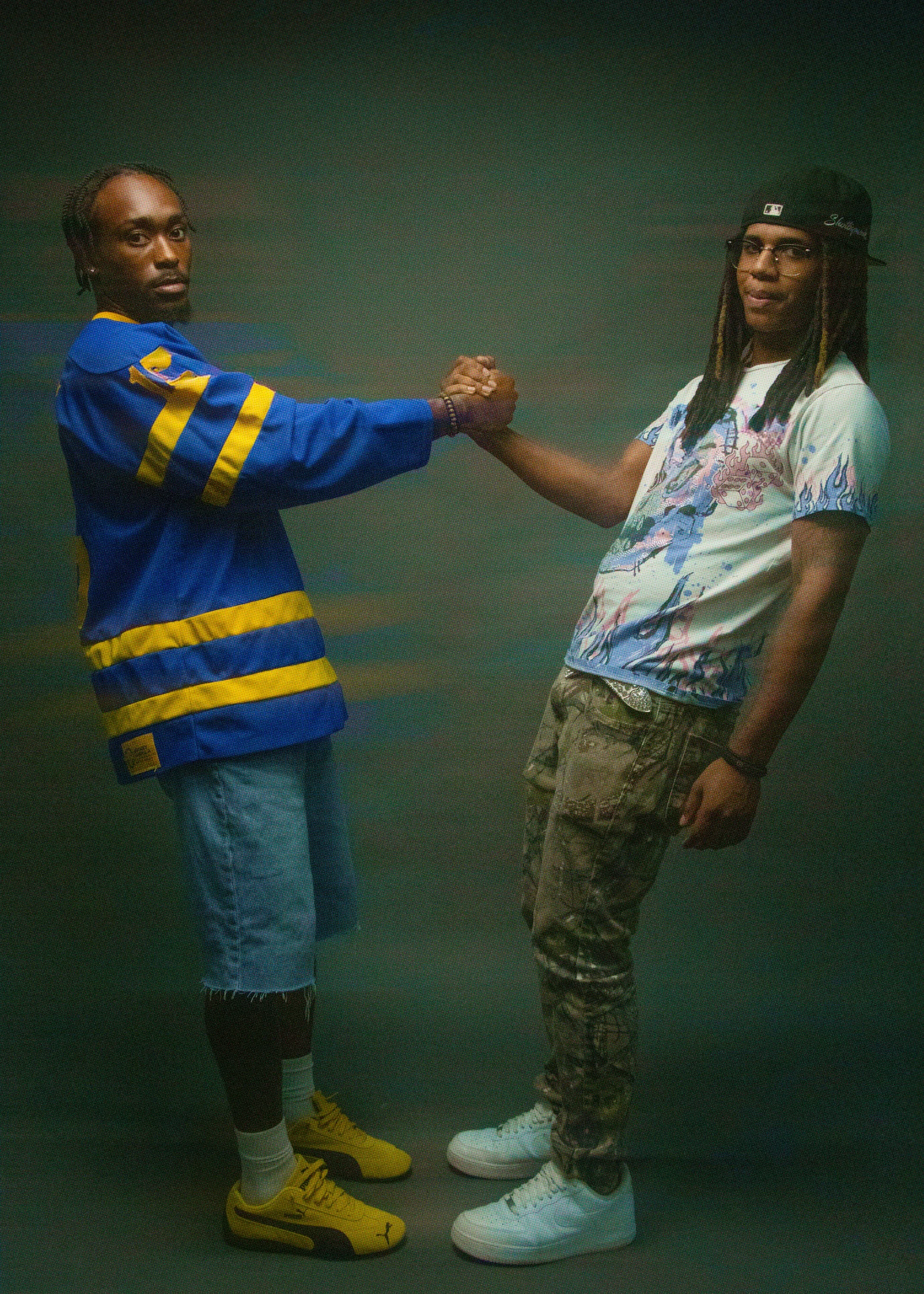 Two young men shaking hands, one dressed in a blue and yellow sports jersey, the other in a graphic t-shirt, camouflage pants, and wearing glasses and a baseball cap, standing against a dark background.