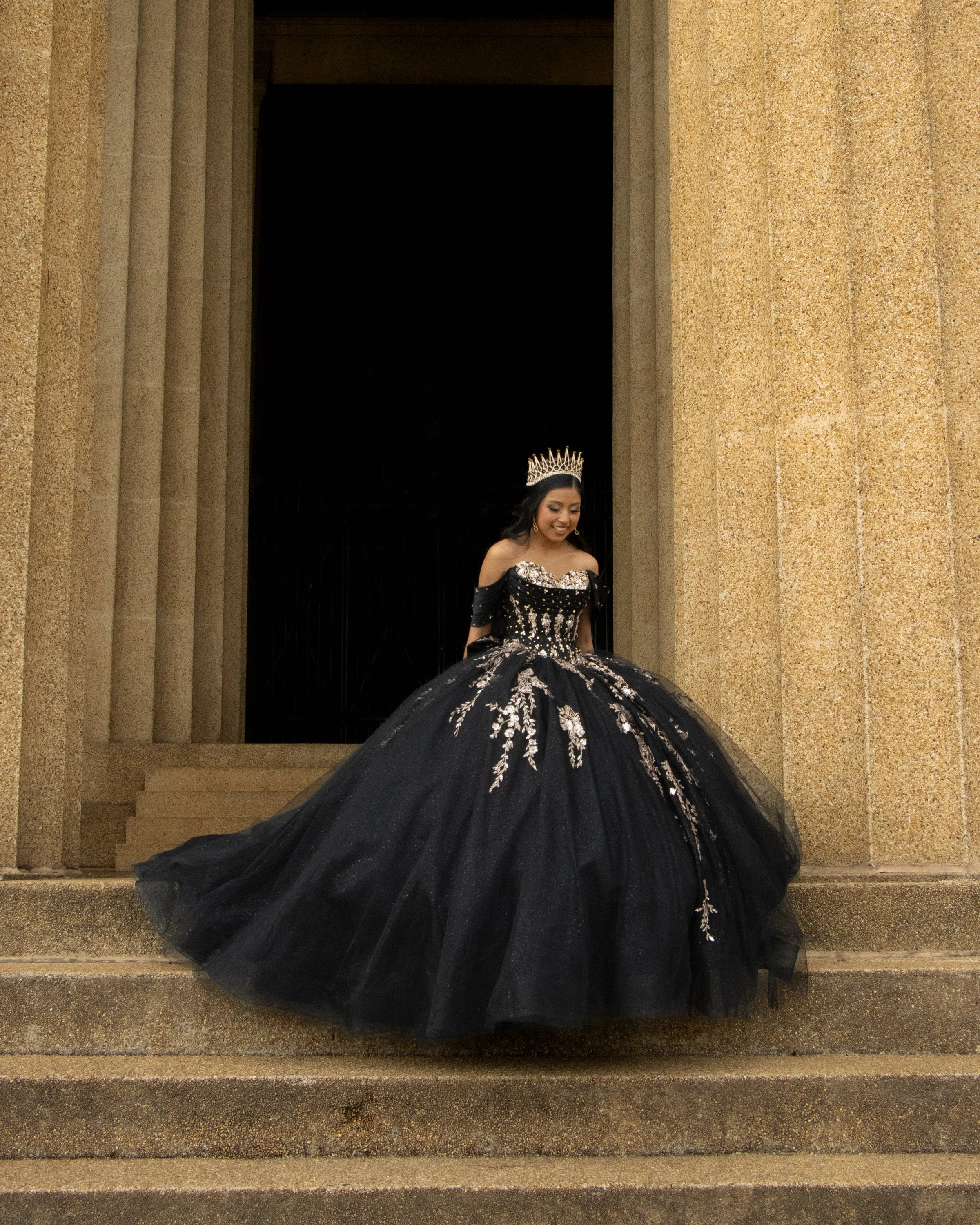 Young woman wearing a crown and a black and silver gown, smiling, standing in front of a large stone building with columns.