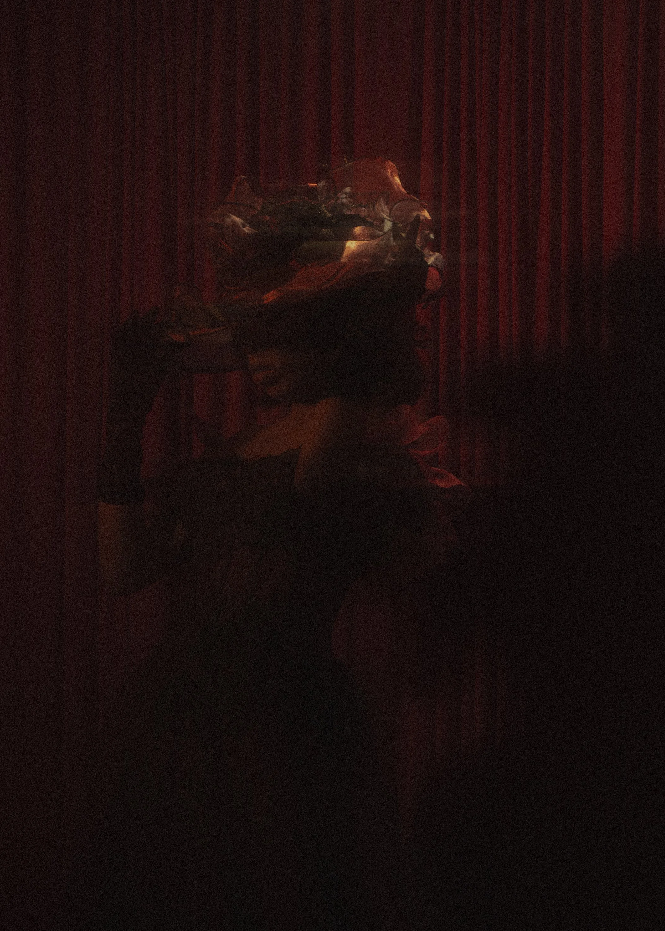 A dark scene with a twisted, reflective sculpture lit by a red light, set against a red curtain background.