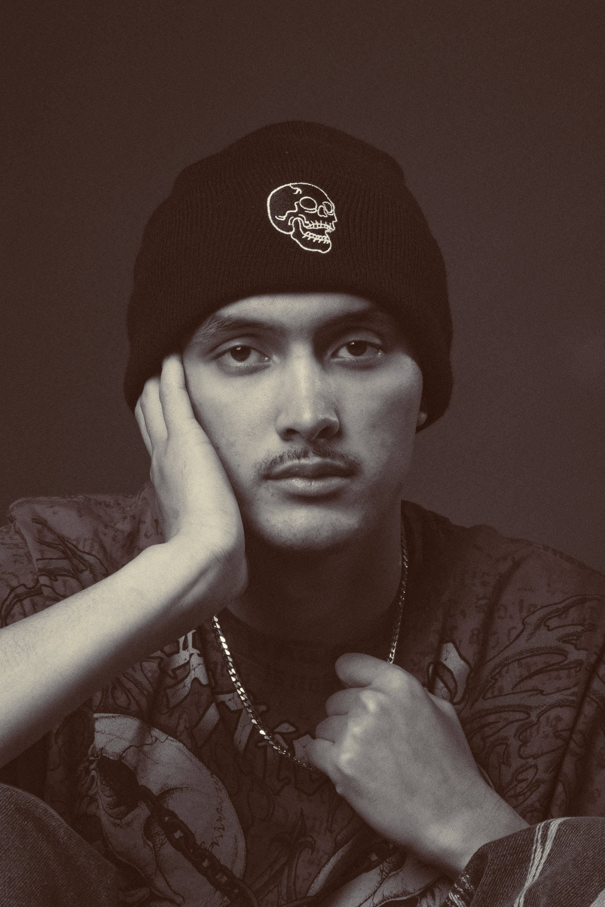 A black and white portrait of a young man wearing a black beanie with a white skull embroidery, resting his face on his left hand and wearing a chain necklace.