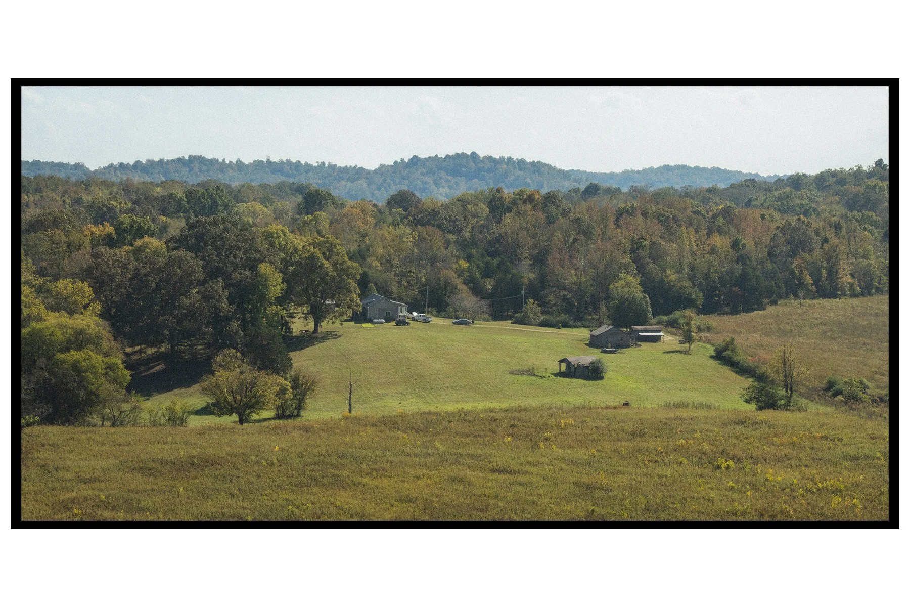 A rural landscape with green fields, trees, houses, and a backdrop of forested hills and mountains under a clear sky.