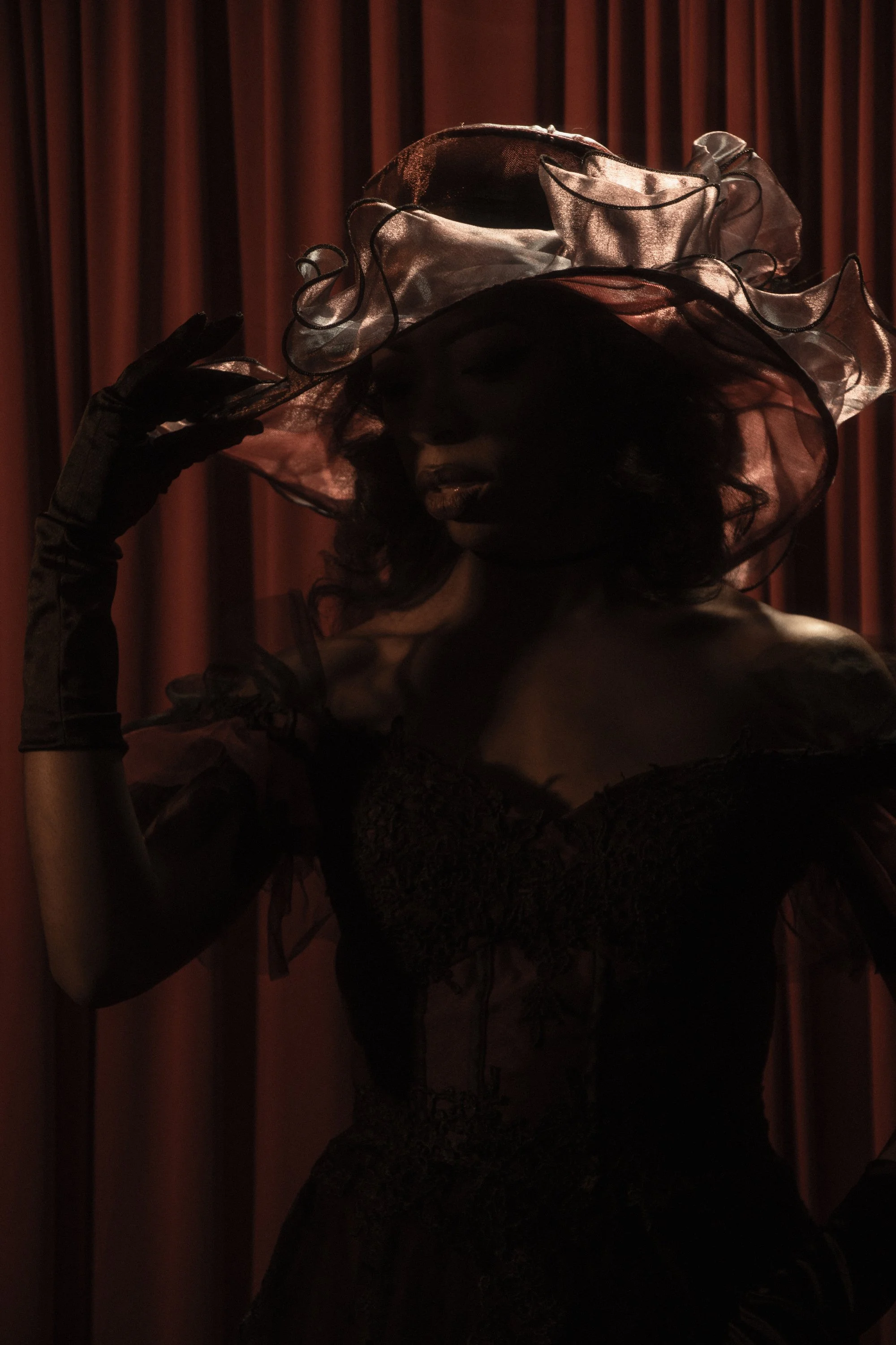 A woman with dark skin wearing a large translucent hat and gloves, touching the brim of her hat, standing in front of a red curtain.