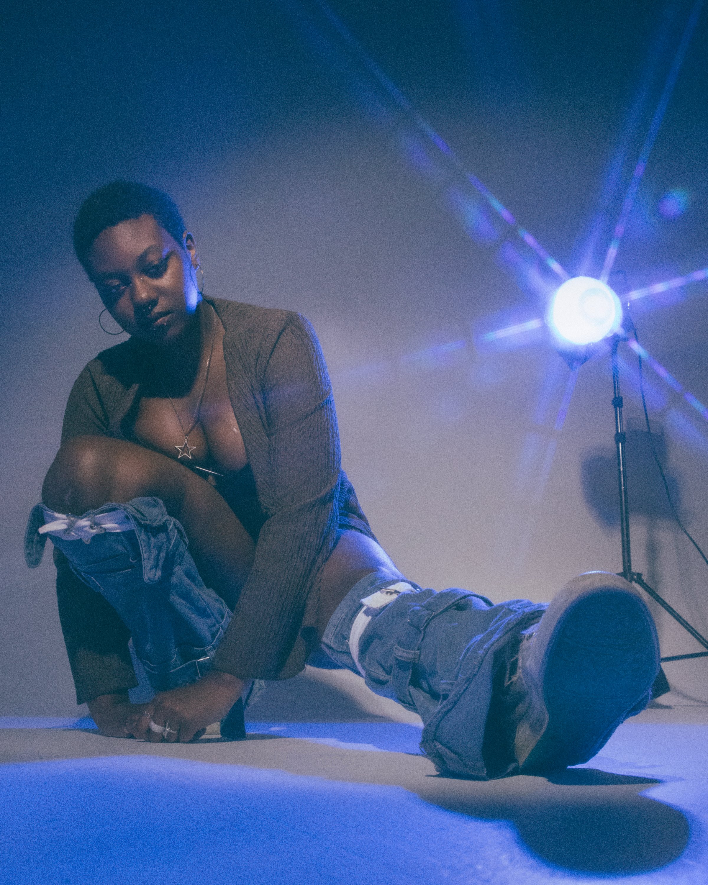 A person with short hair and hoop earrings, wearing a necklace, sits on the floor in a dimly lit space with blue lighting and a bright light source on a stand to the right.