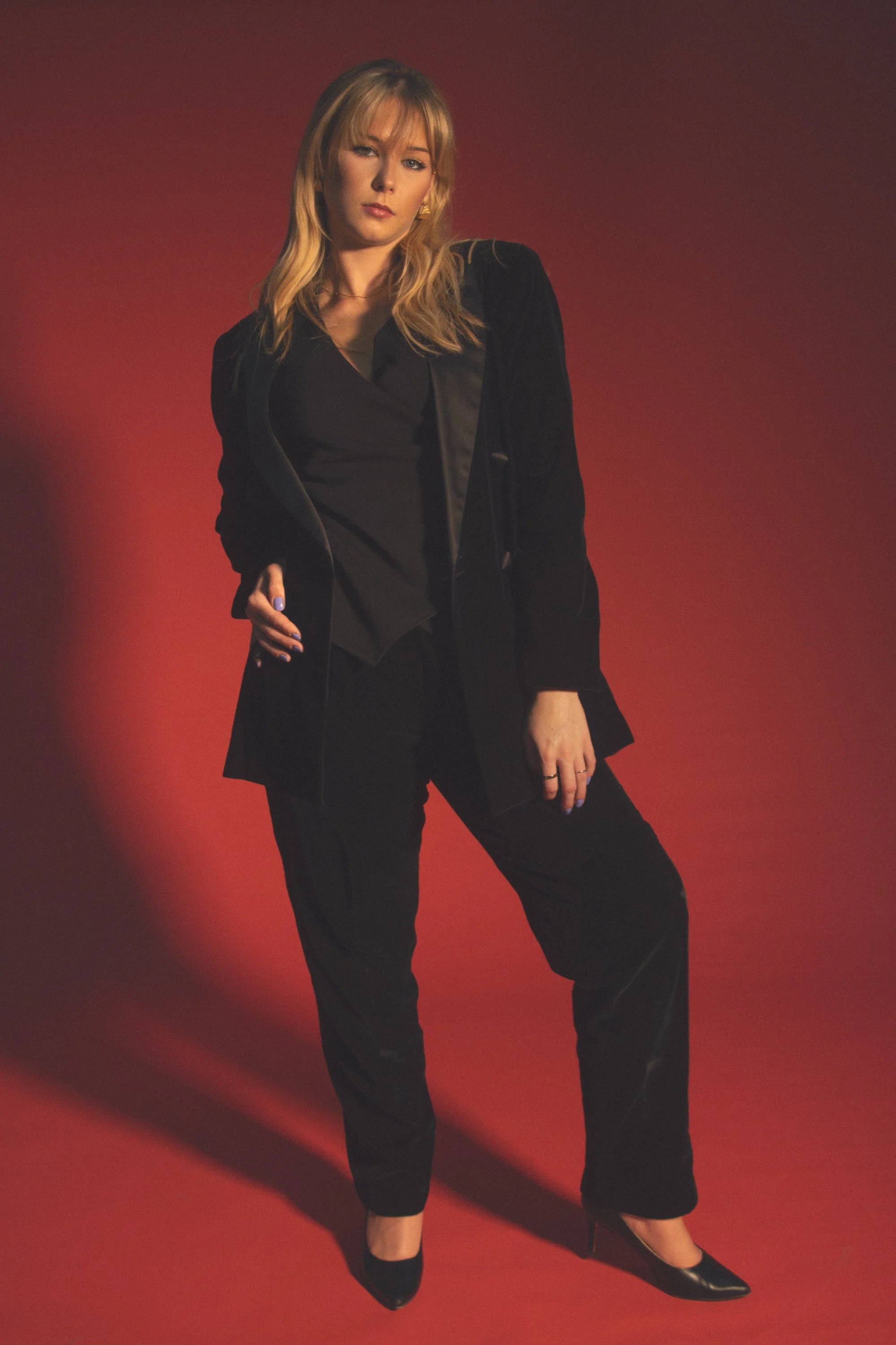 A woman with blonde hair wearing a black blazer, black top, black pants, and black pointed shoes posing against a red background.