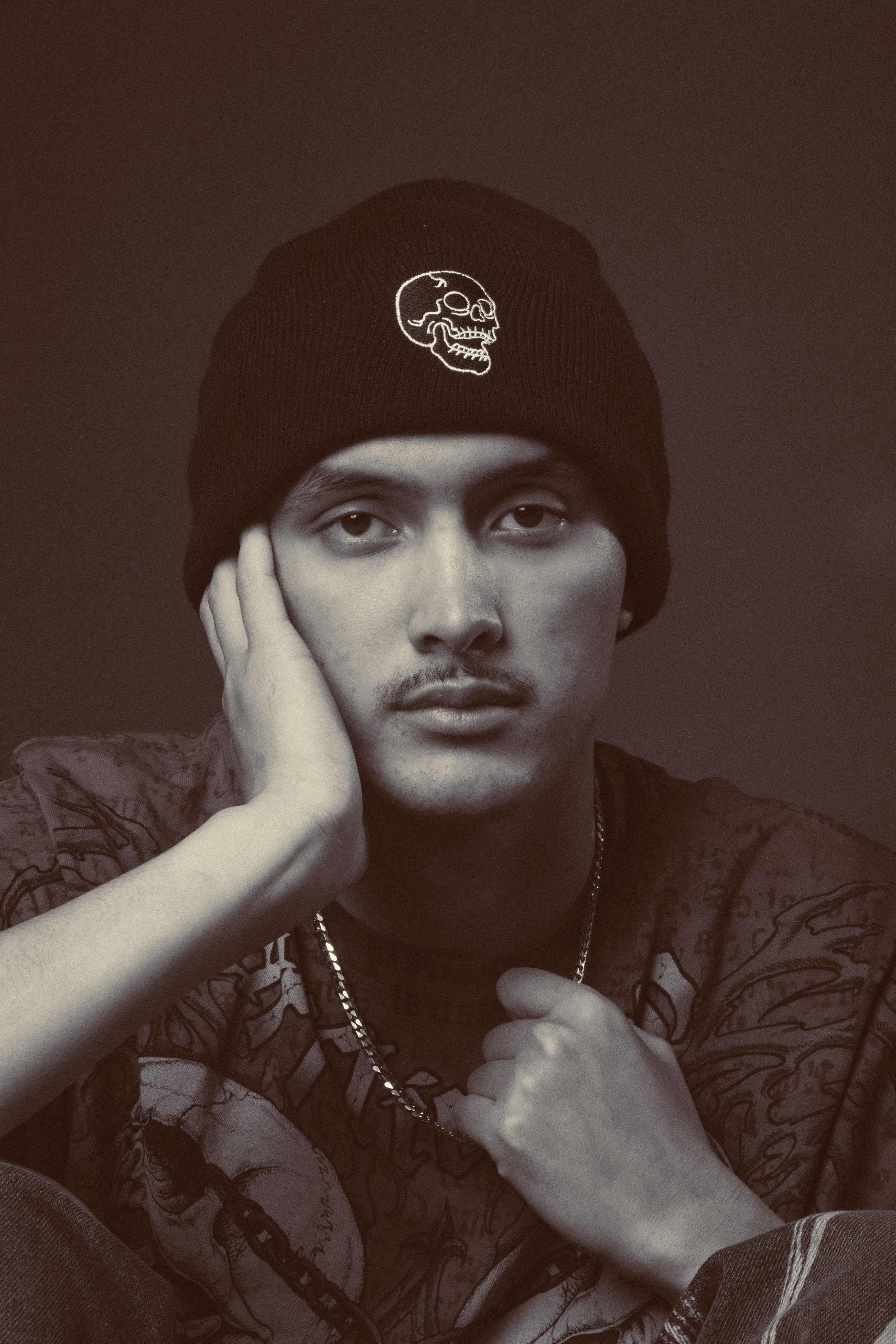 Portrait of a man wearing a black beanie with a skull design, resting his head on his hand, looking directly at the camera in a black-and-white photograph.