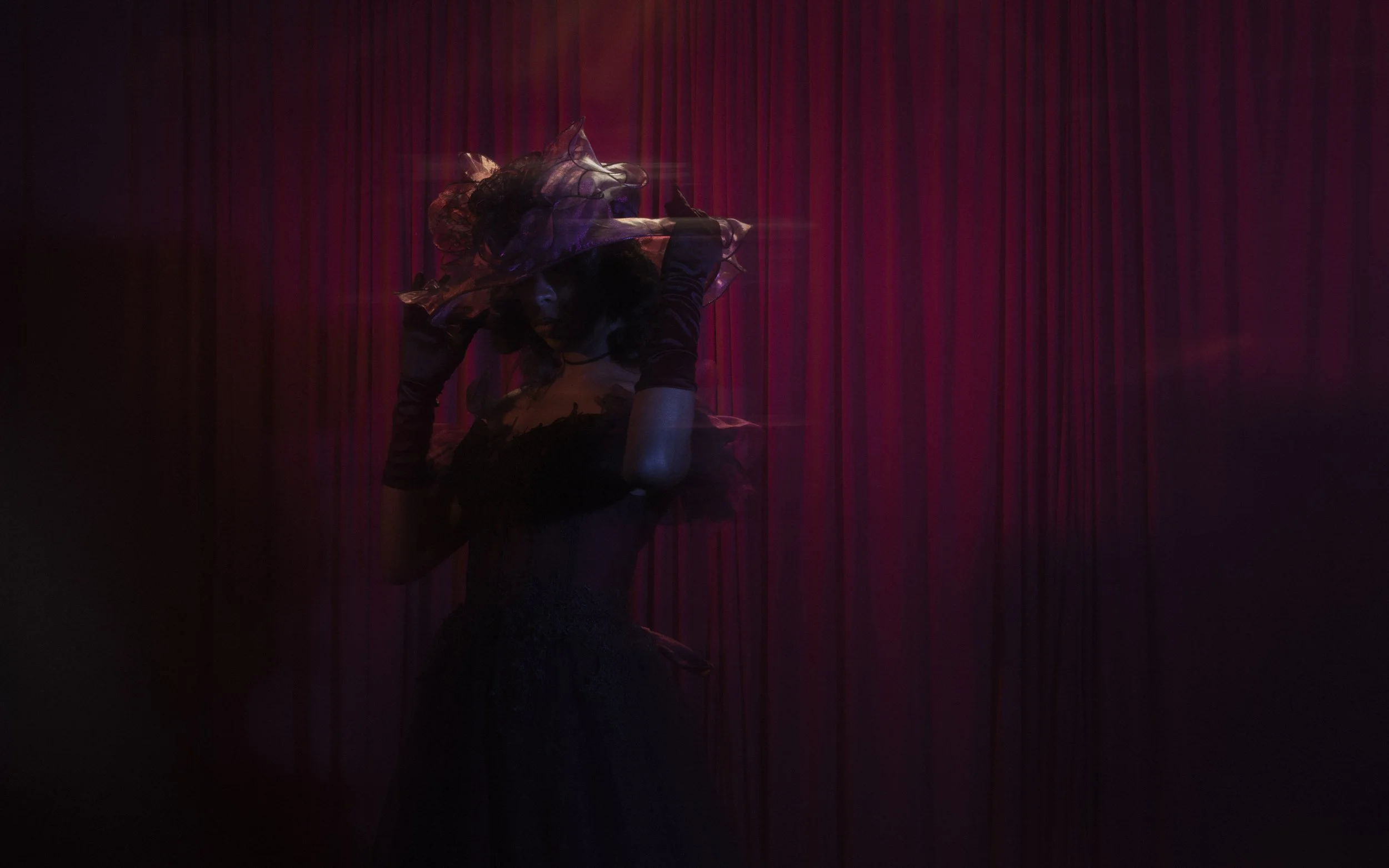 A person dressed in dark clothing and a hat with mesh overlay, posed on a stage with red curtains in the background, under low lighting.