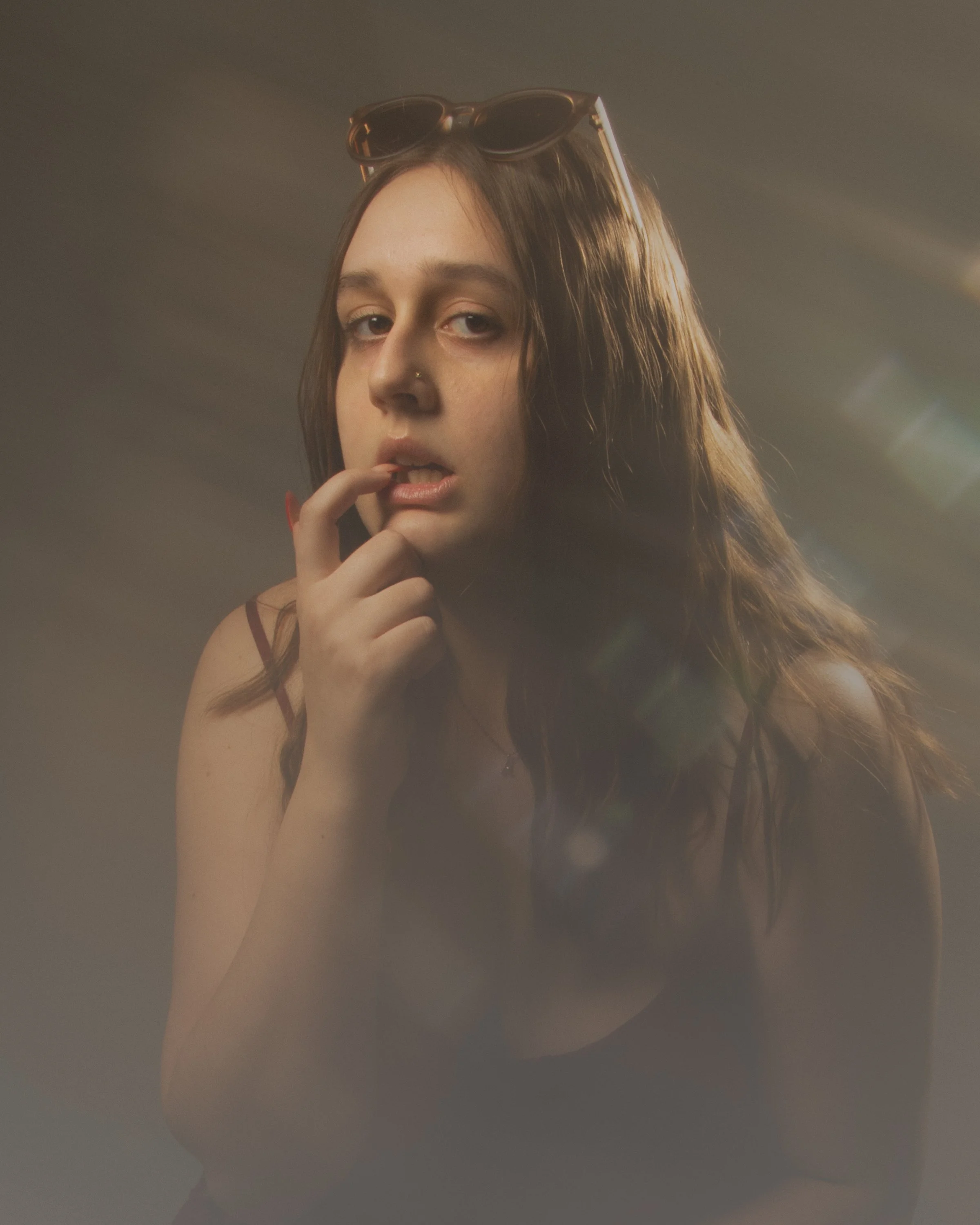 A young woman with long brown hair, sunglasses on her head, and a nose piercing touching her lips while looking at the camera.