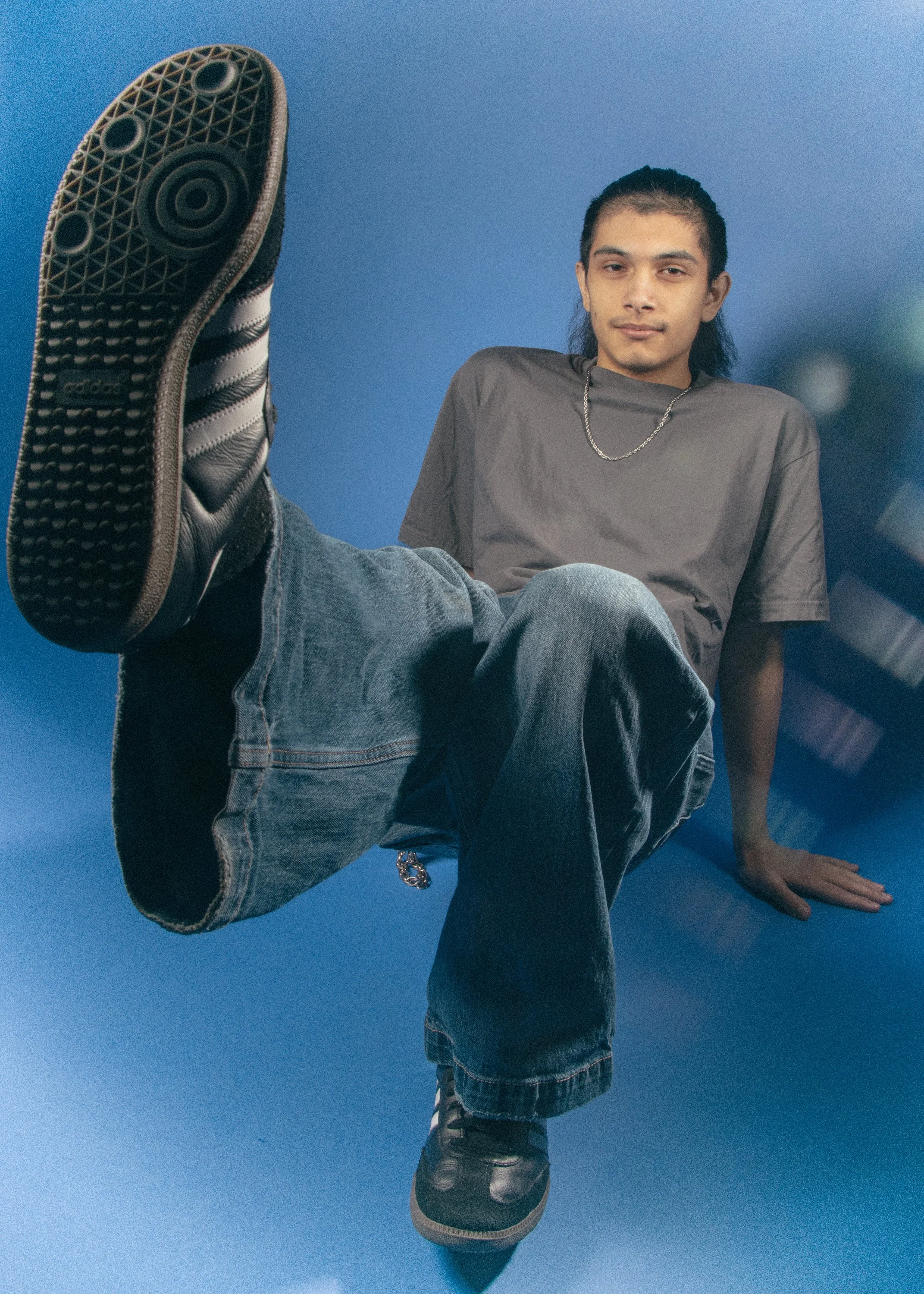 A person with long hair wearing a gray T-shirt, jeans, and black sneakers, seated with one leg extended toward the camera, against a solid blue background.