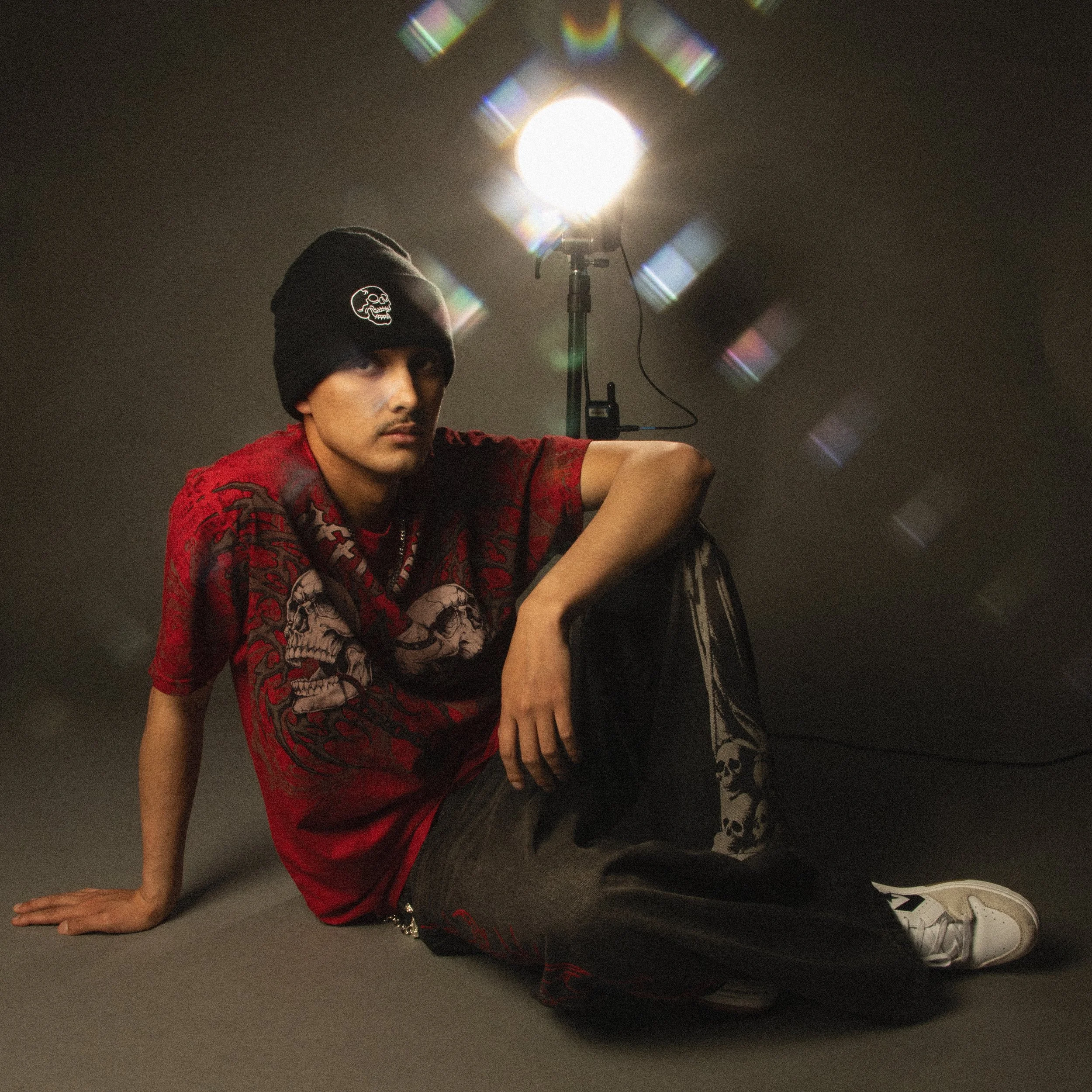 A person wearing a black beanie with a skullnylon design, a red graphic t-shirt, and white sneakers, sitting on the floor next to a professional photography or filming light.