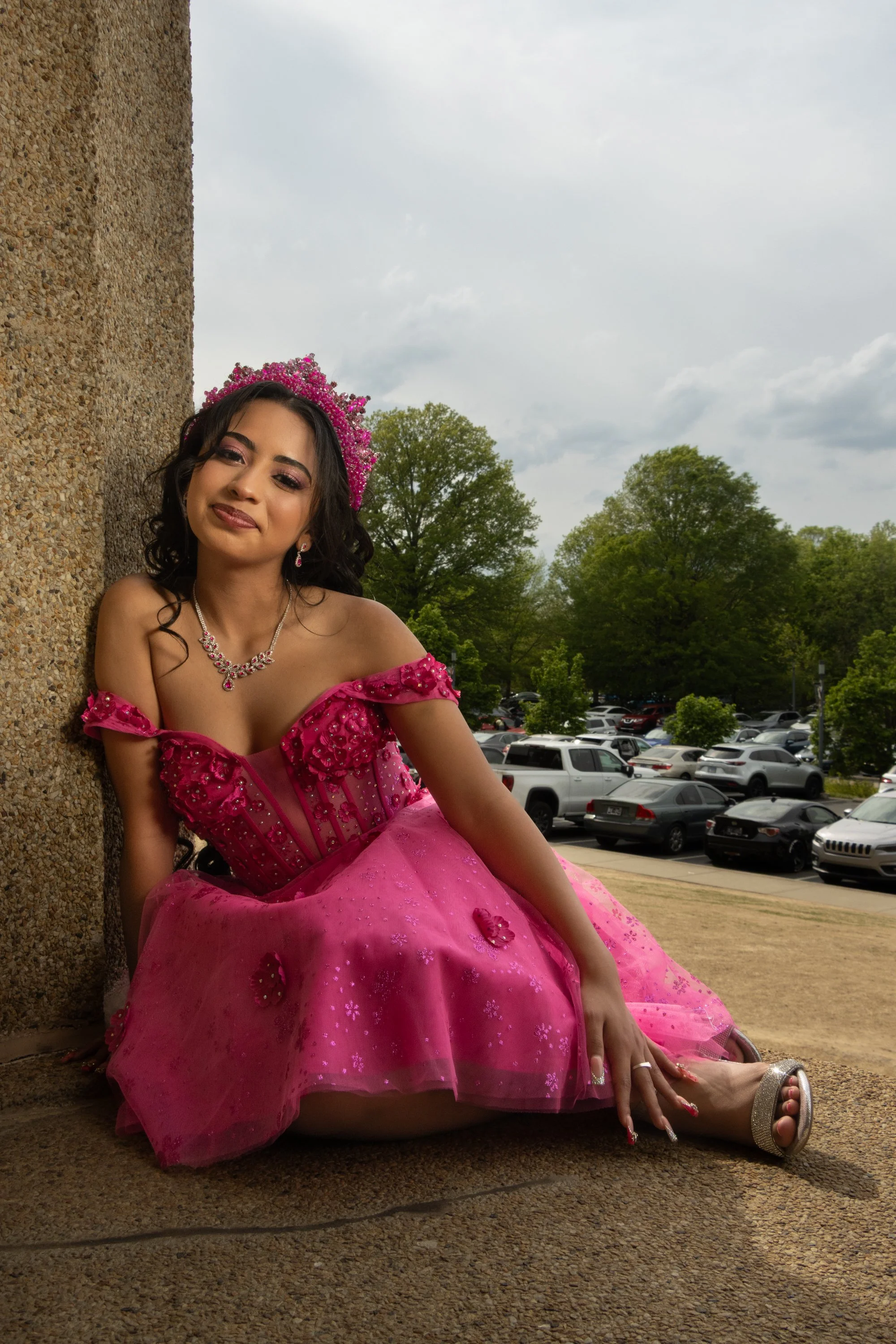Young woman in a pink dress with jewelry and a tiara, sitting against a wall outdoors, with trees and a parking lot in the background, overcast sky.