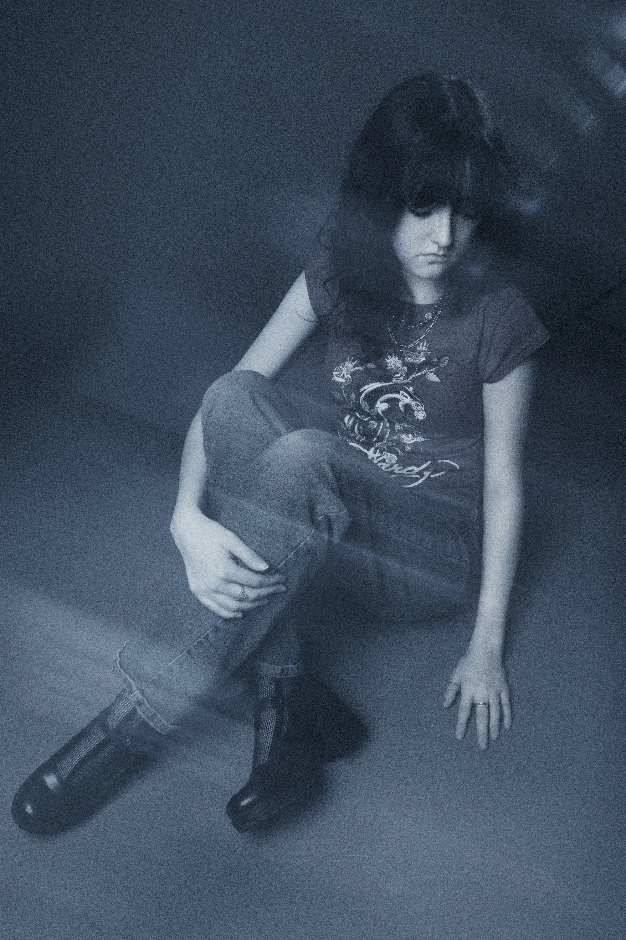 A woman with curly hair, seated, wearing a graphic t-shirt, jeans, and boots, with her hand resting on her knee in a dimly lit room.