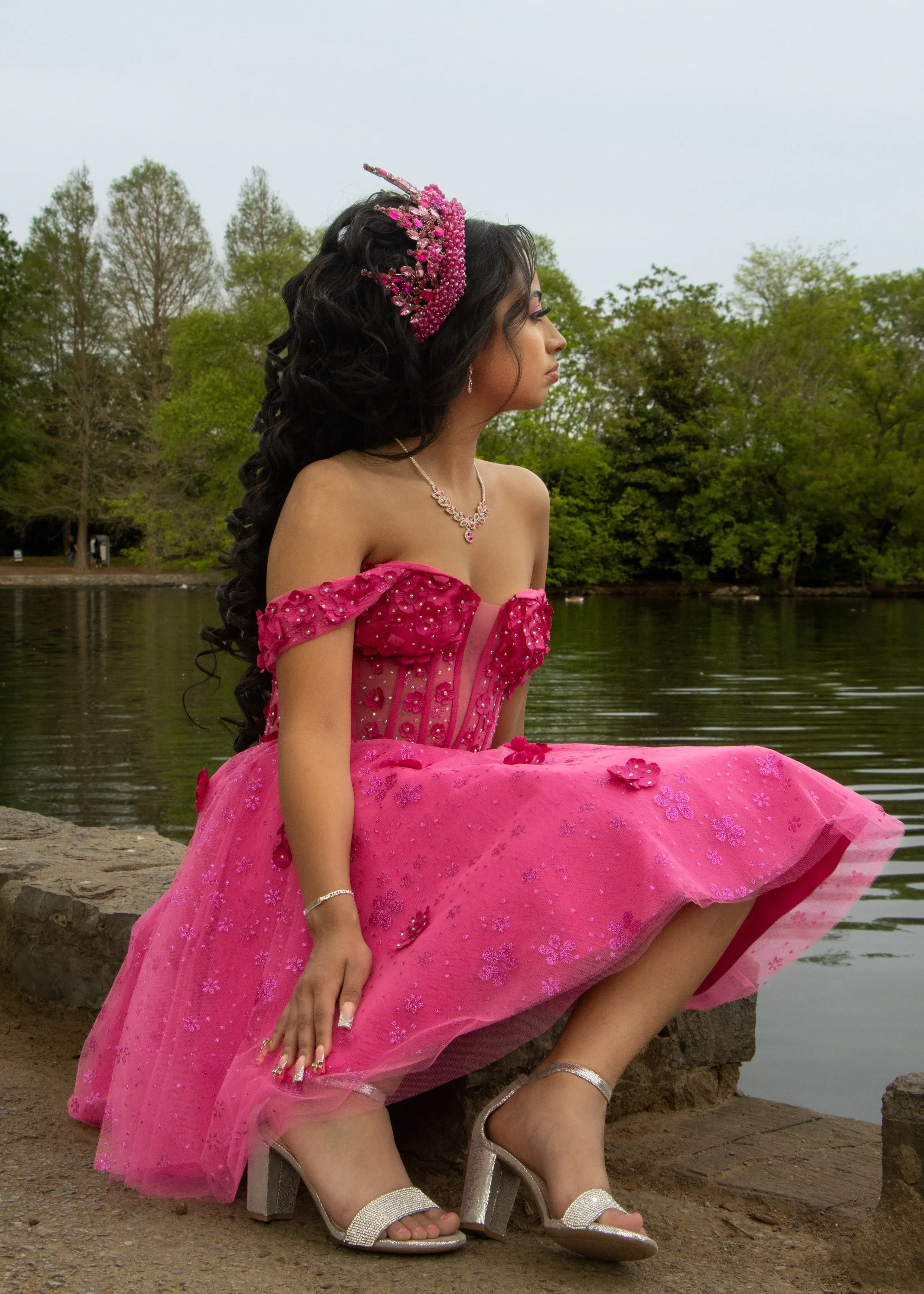 Young woman in a pink ball gown sitting outdoors near a lake or river with trees in the background, wearing high-heeled sandals, jewelry, and a tiara.
