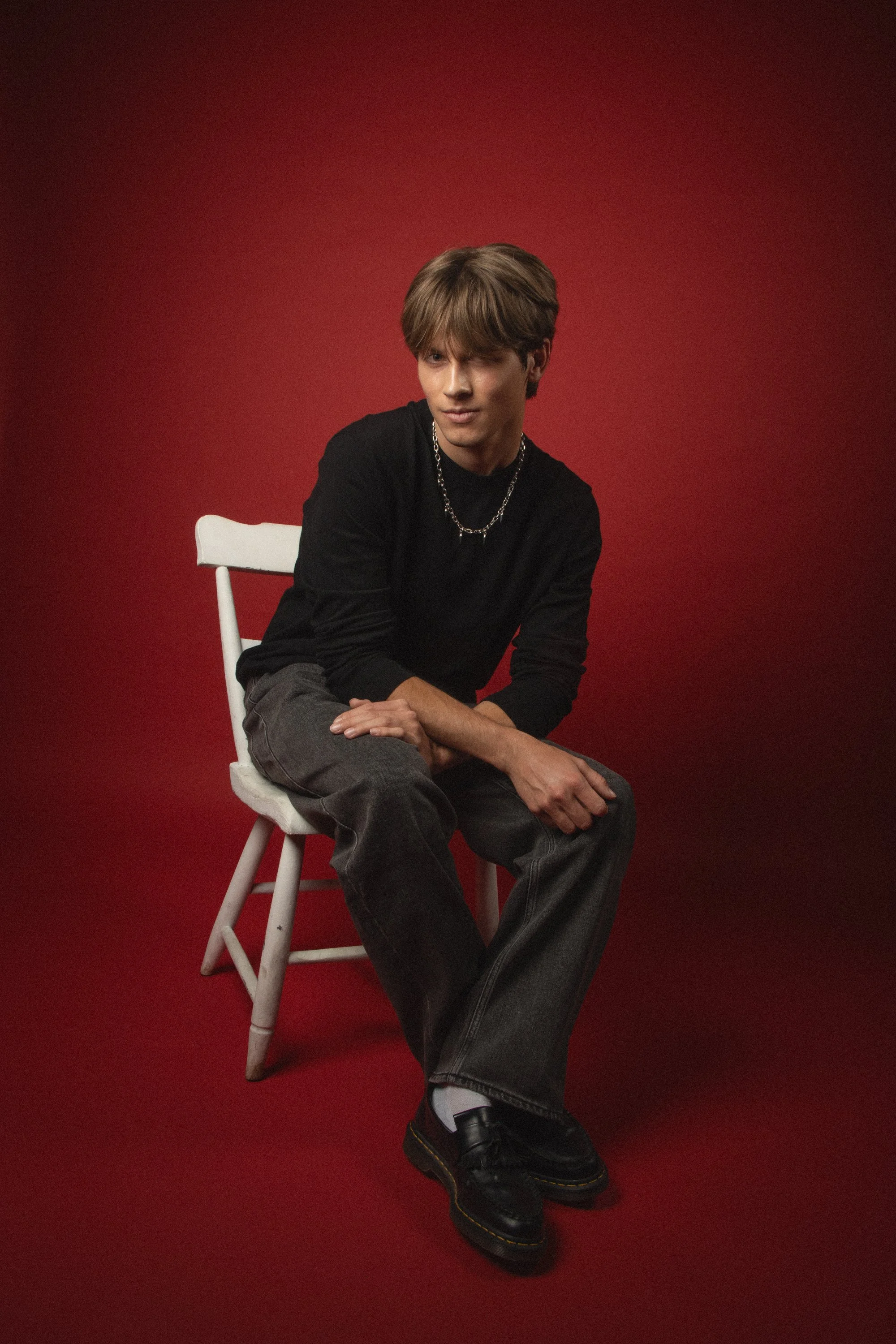 A young man with light brown hair sitting on a white chair against a red background. He is wearing a black long-sleeve shirt, gray jeans, black shoes, a silver chain, and a ring. His legs are crossed, and his hands are resting on his lap.