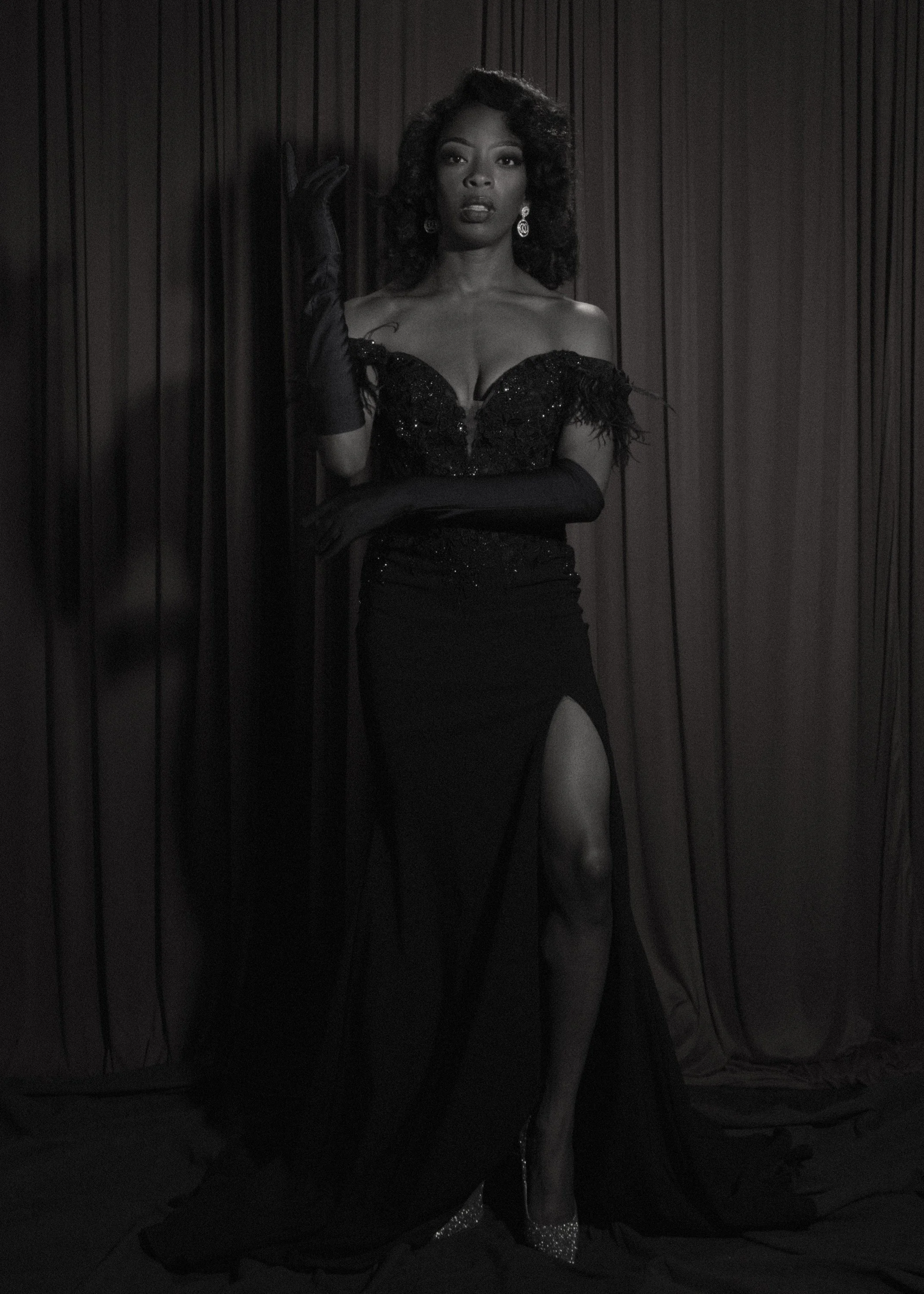 A woman standing in front of a dark curtain, wearing an off-shoulder black dress with a high slit, long black gloves, and sparkling high heels.