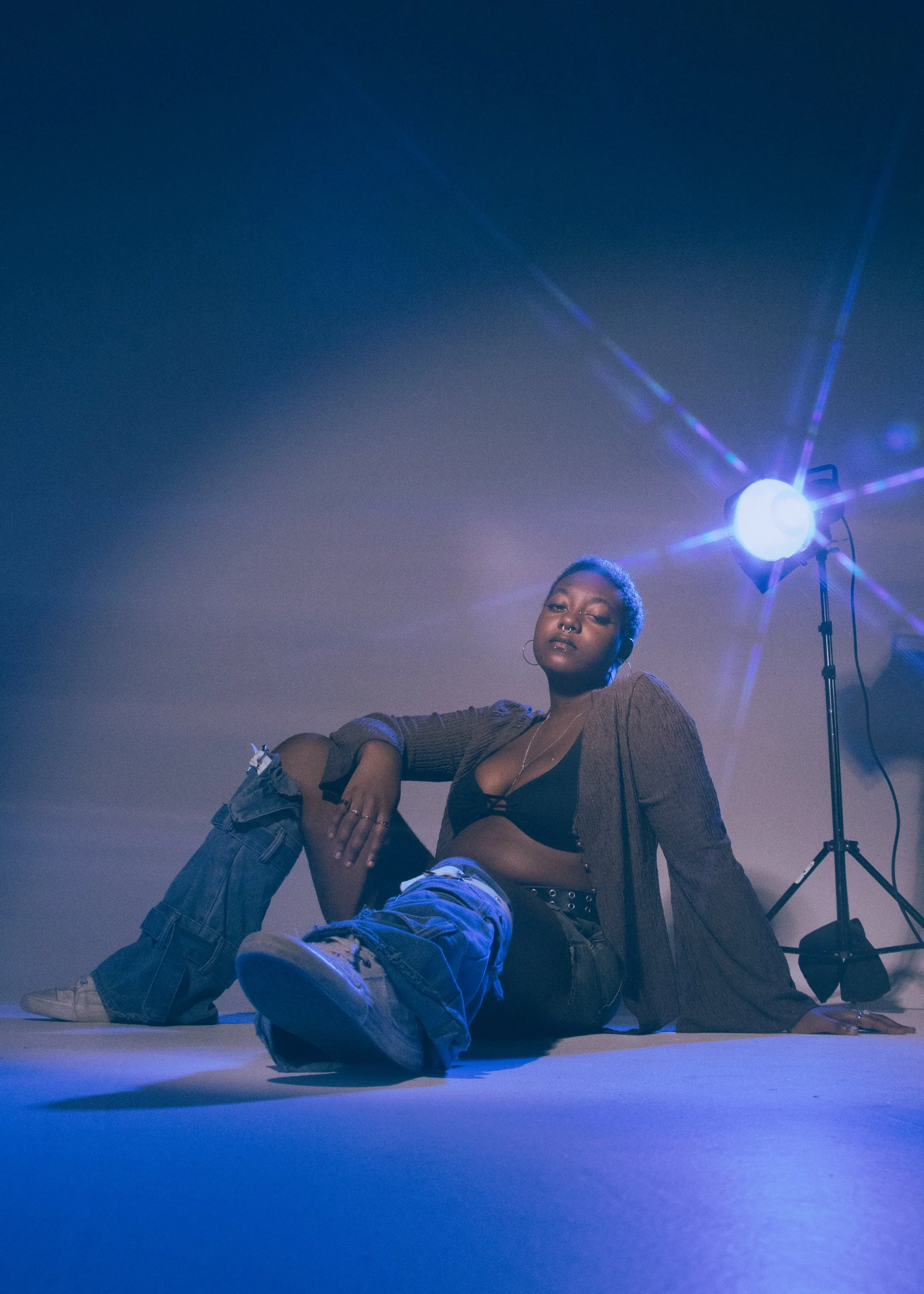 A woman sitting on the floor with her legs crossed, wearing a brown jacket, black top, and ripped jeans, posing in front of a bright blue light in a photography studio, with a neutral expression.