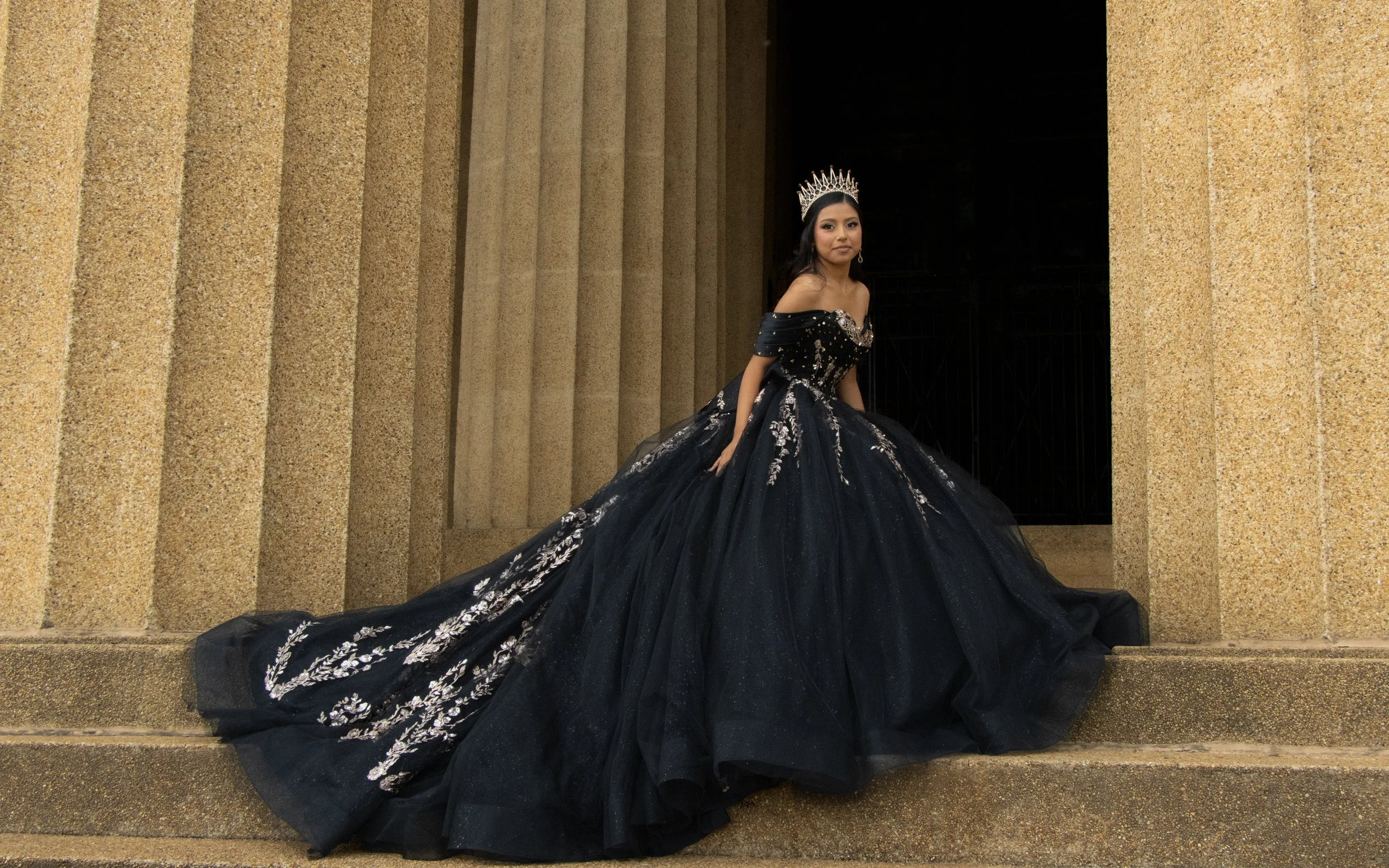 A young woman in an off-shoulder black gown with intricate silver embroidery, wearing a silver crown, stands on steps between large stone pillars.