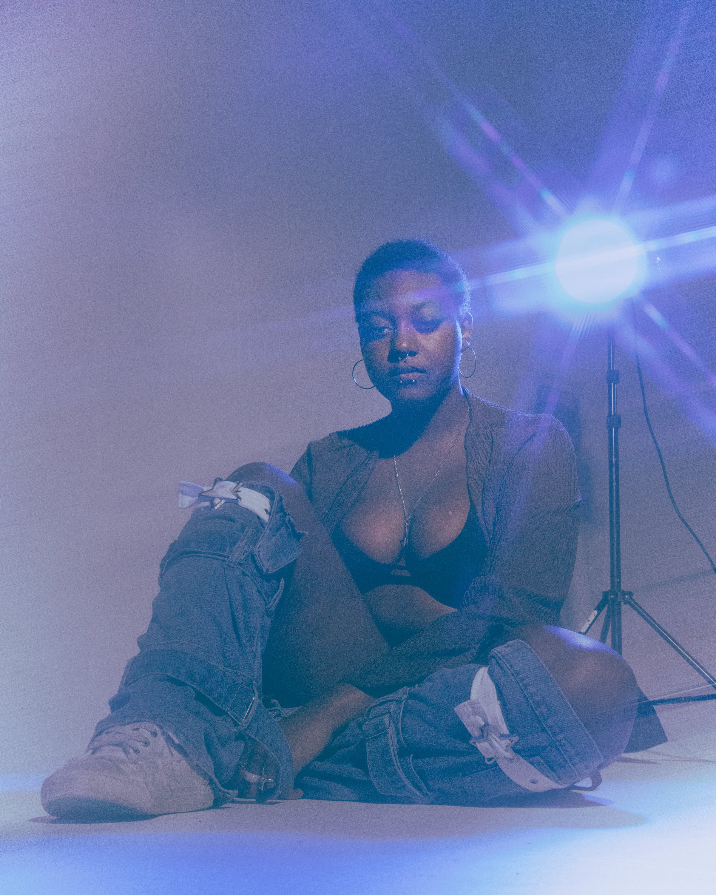 A person with short hair and earrings sitting on the floor in front of a bright studio light with purple and blue hues, wearing a texture top and sneakers.