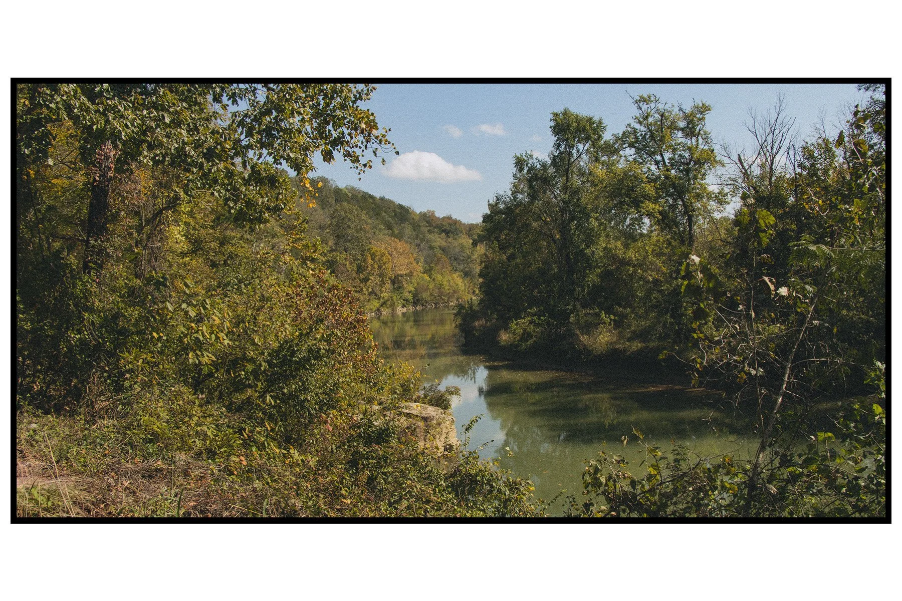 A peaceful river flowing through a lush, green forest with trees showing fall foliage and a clear blue sky with a few clouds overhead.