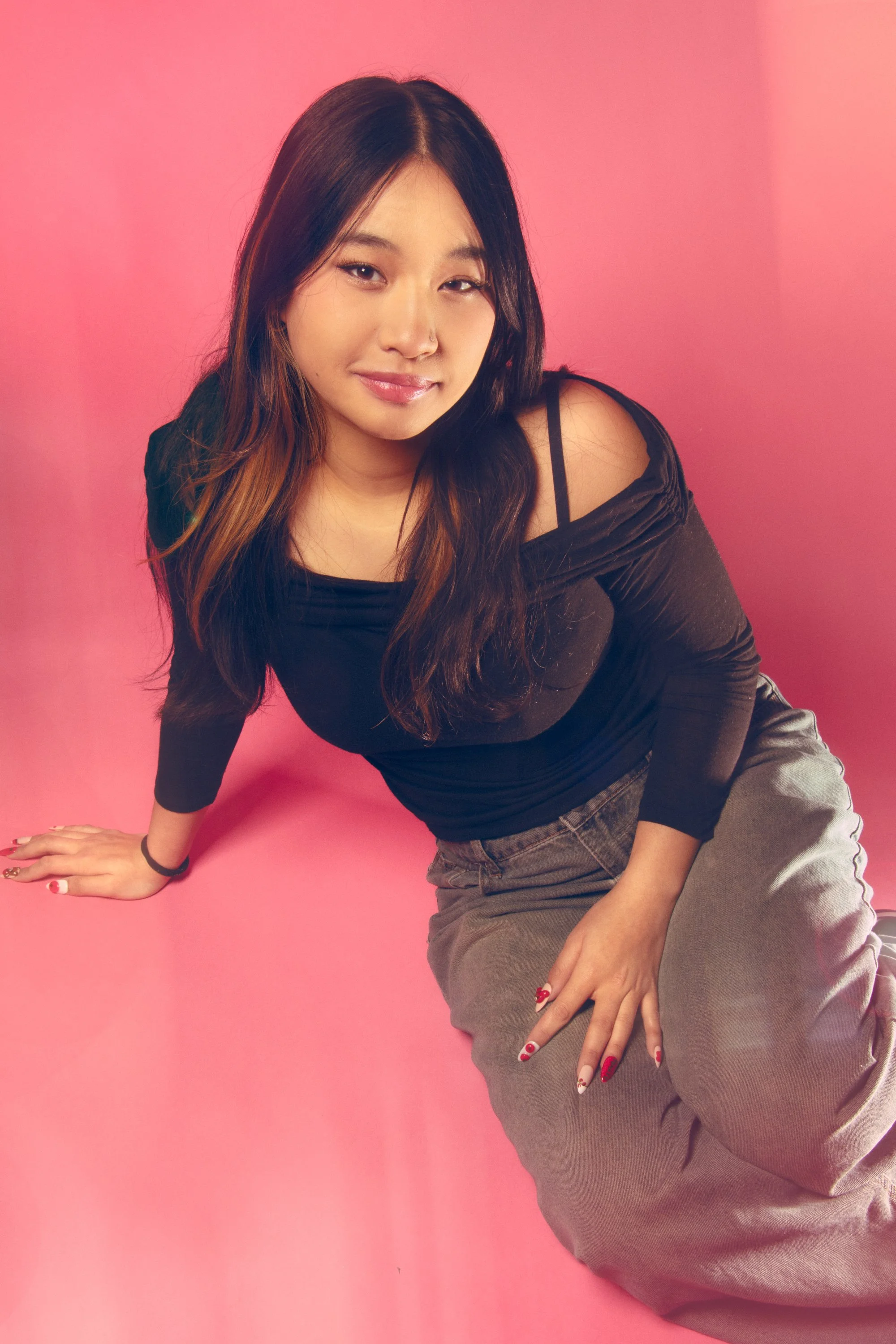 A young woman with long dark hair, wearing a black off-shoulder top and gray pants, sitting against a pink background, smiling lightly.