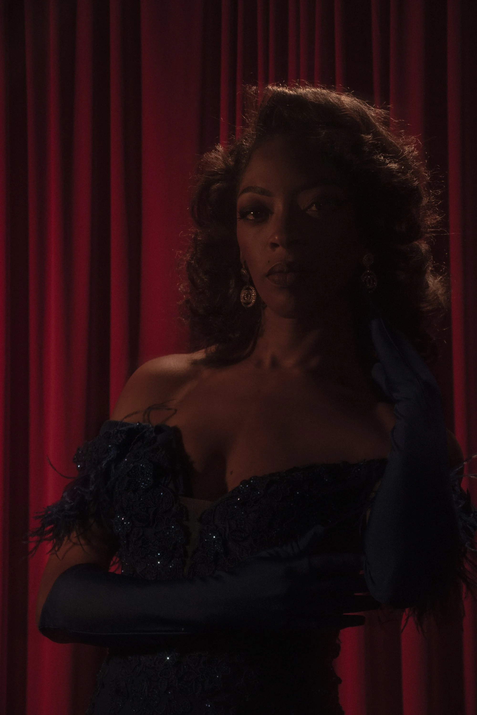 A woman with curly hair and dark makeup, wearing earrings, dressed in a dark, ornate outfit with lace and ruffles, standing in front of red curtains.