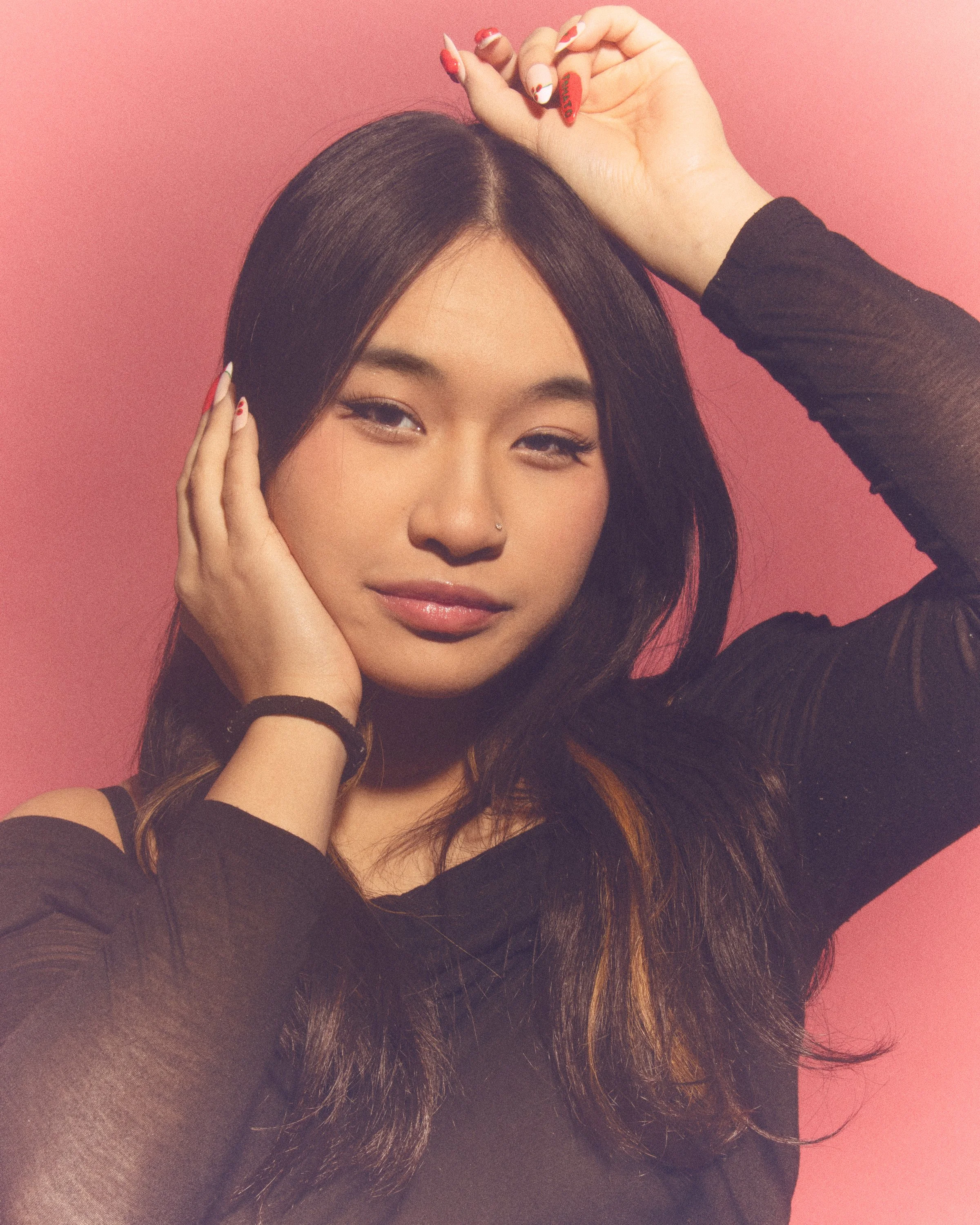 A woman with long, dark hair and a nose piercing touches her face with her left hand and raises her right hand above her head, against a pink background. She has styled nails with red and white designs and is wearing a black, sheer, long-sleeved top.