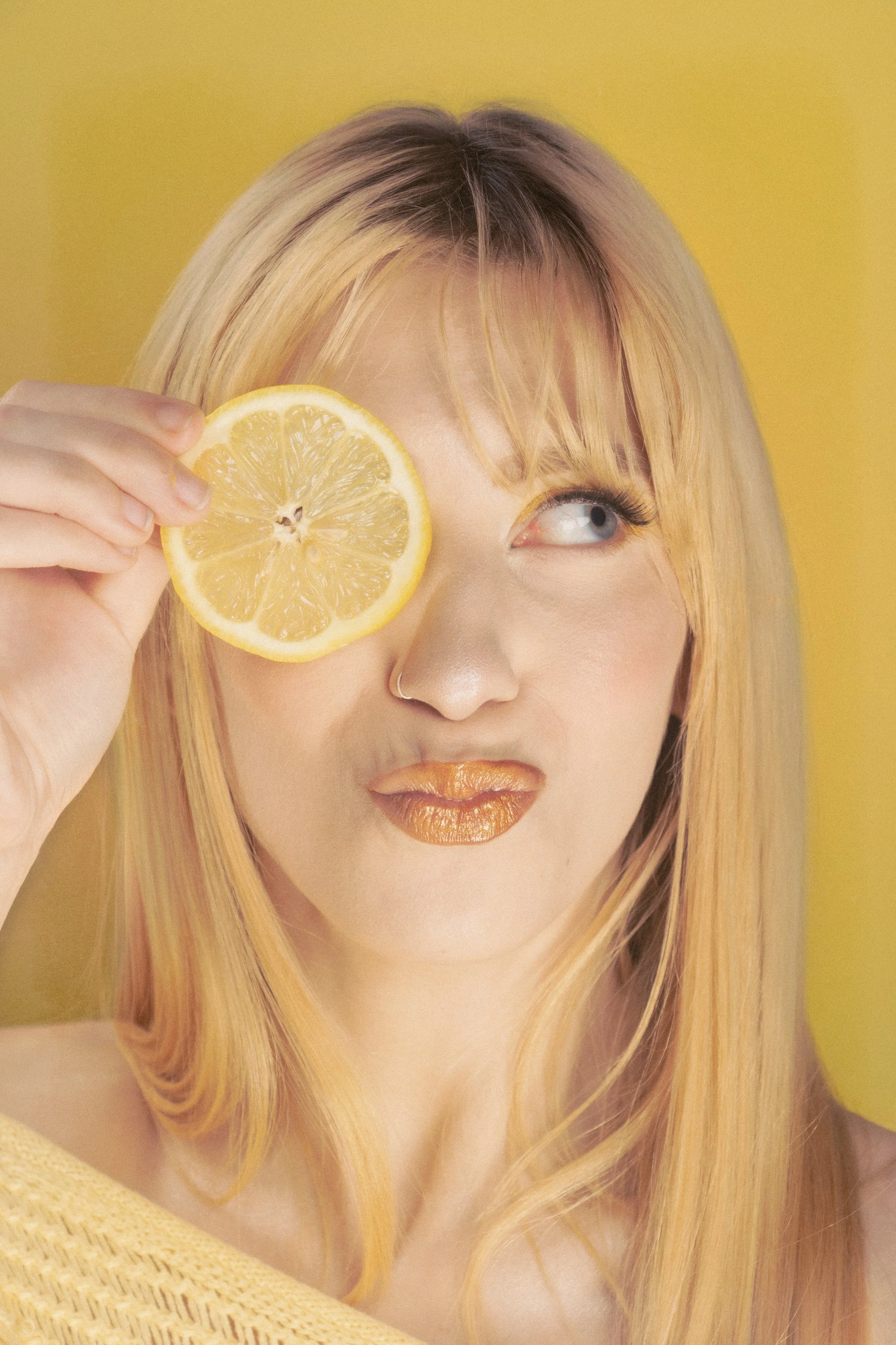 A woman with long blonde hair and blue eyes holds a lemon slice over her right eye. She has gold lipstick, a silver nose ring, and a yellow background.