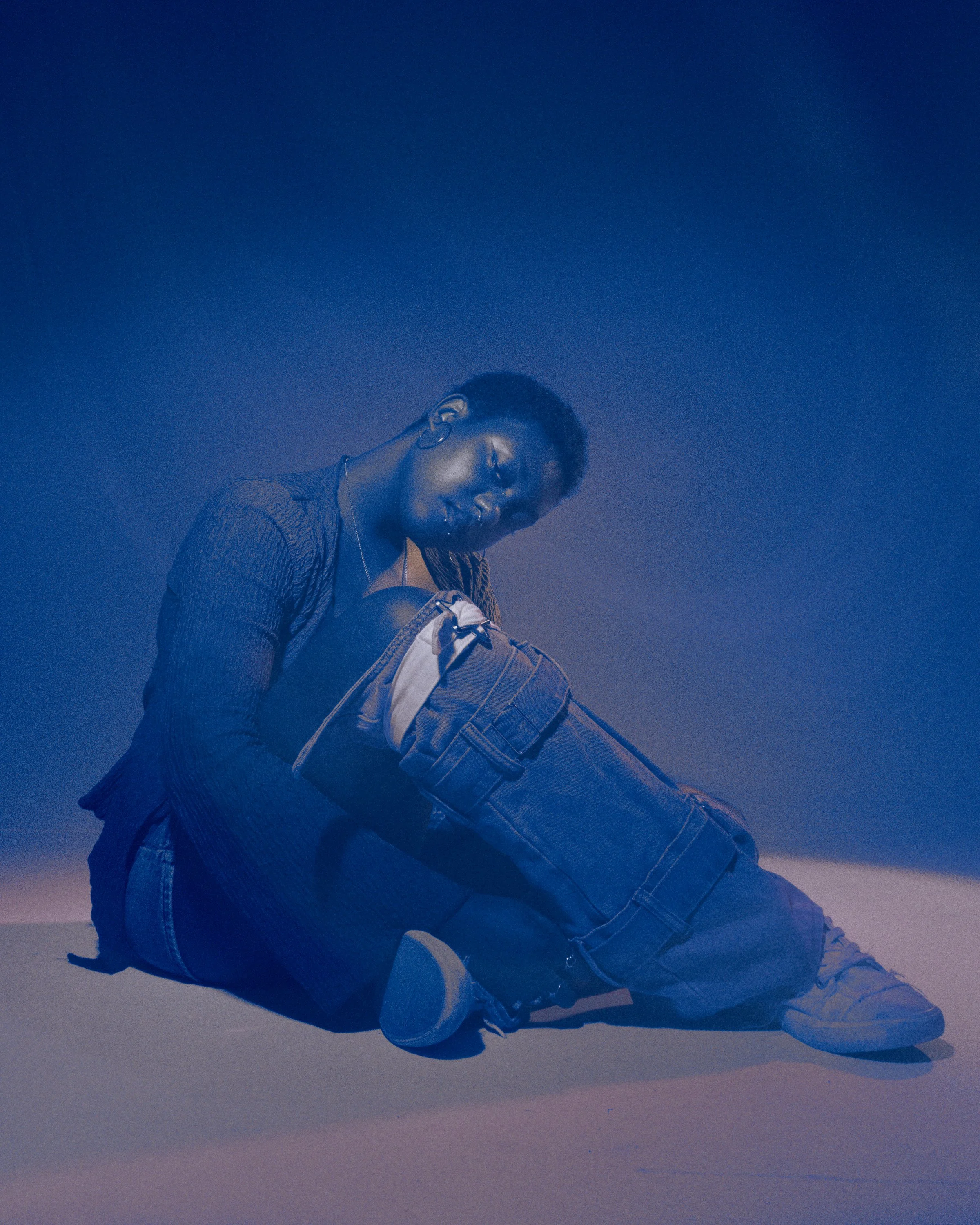 A person wearing a jacket and jeans sitting on a flat surface with a dark sky background, illuminated by blue lighting.