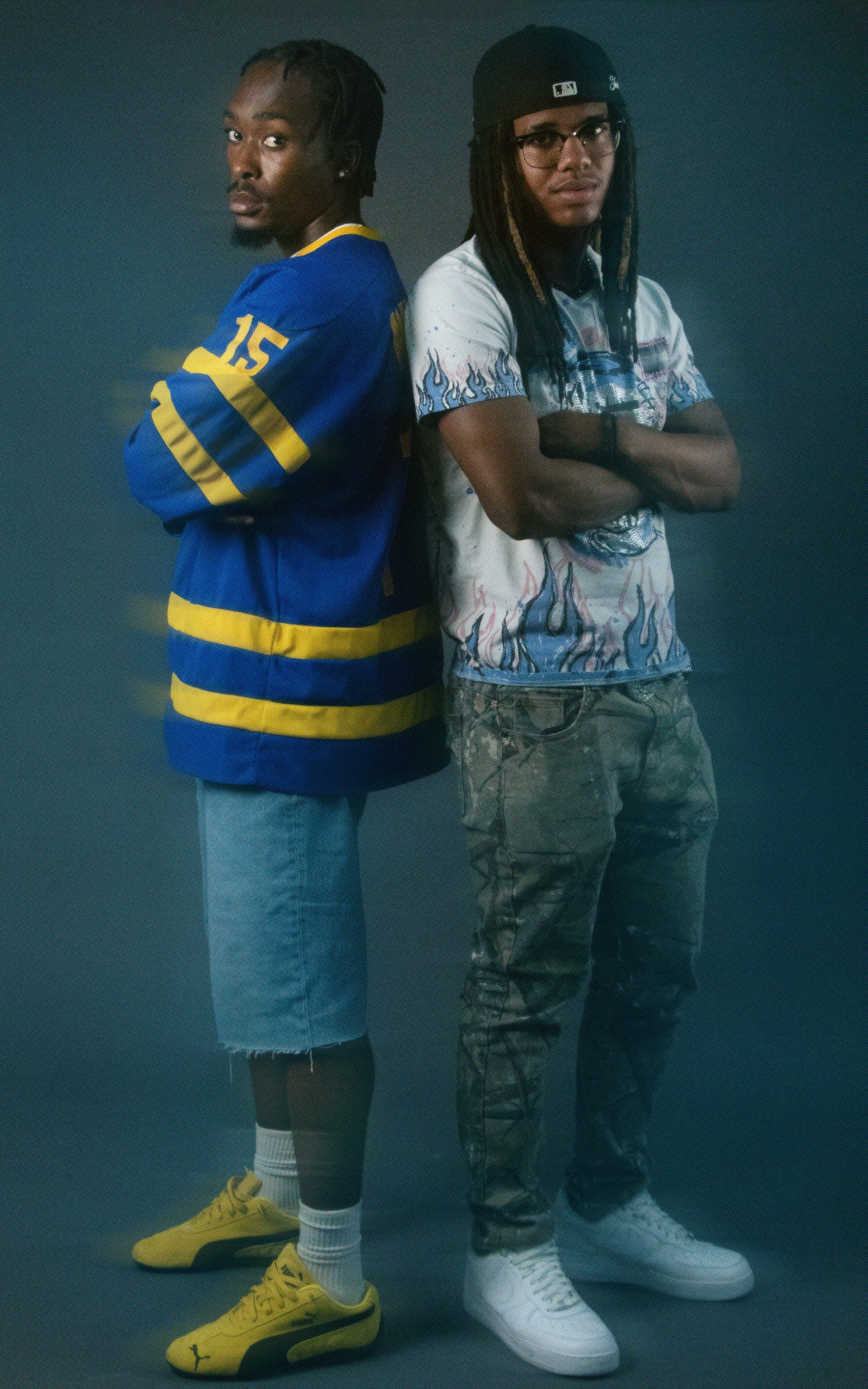 Two young men standing side by side with arms crossed, facing forward. The man on the left is wearing a blue and yellow sports jersey, denim shorts, and yellow sneakers. The man on the right is wearing a graphic t-shirt, camouflage pants, and white s