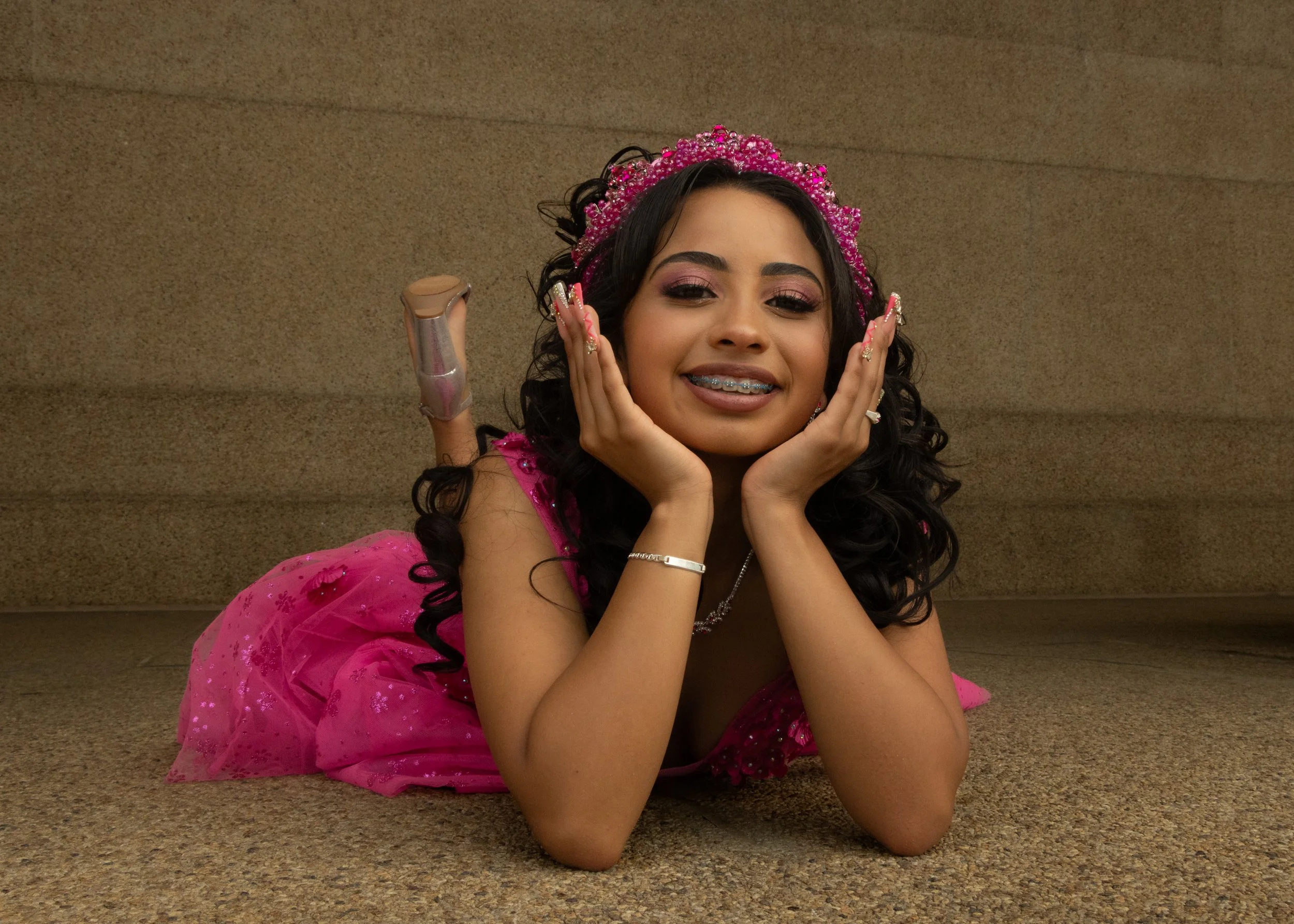 Young woman lying on the floor, wearing a pink dress and a pink beaded tiara, smiling and touching her face, with dark curly hair and braces.