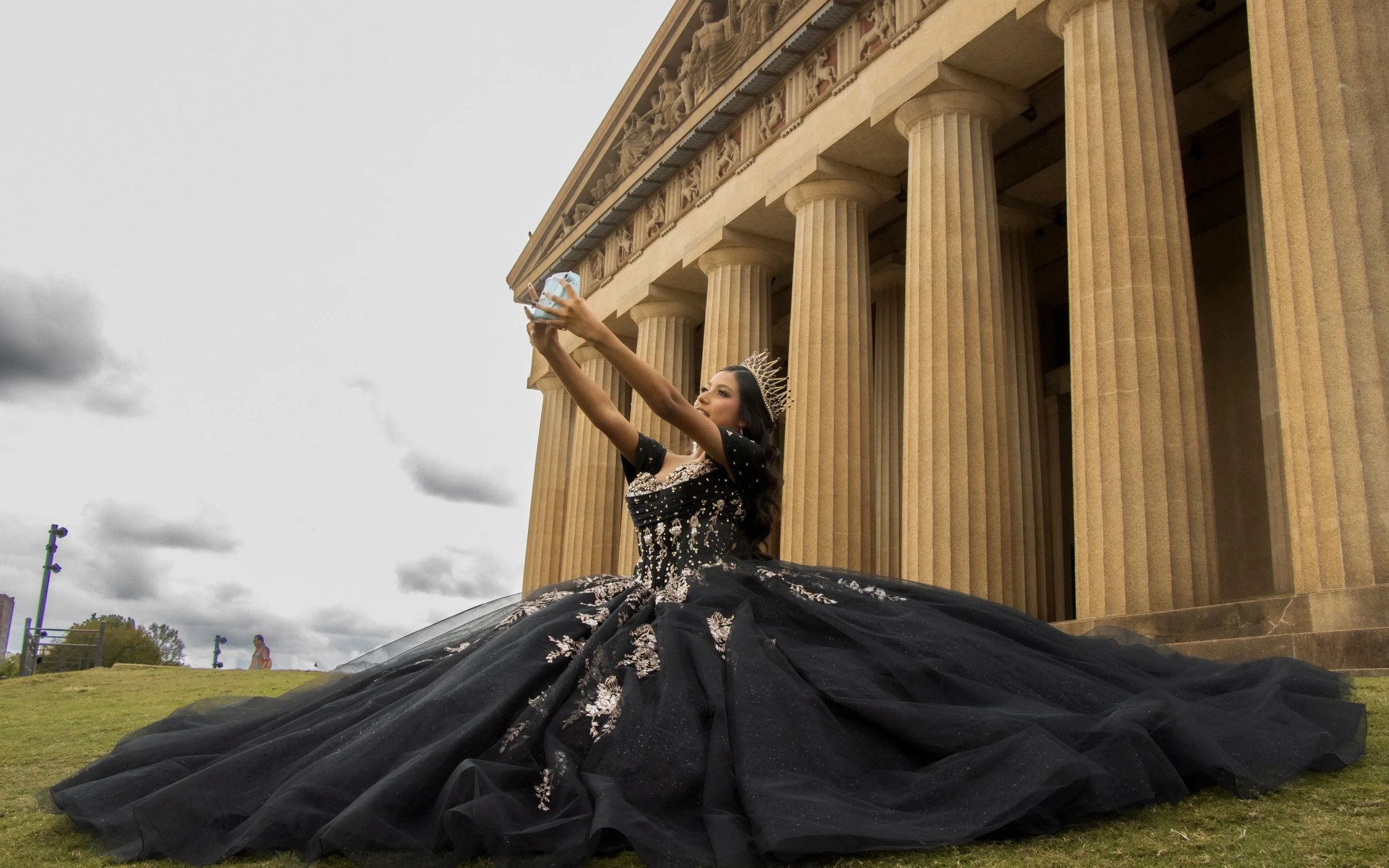 A woman dressed as a queen, wearing a crown and a black and silver gown, sitting on the grass in front of the Lincoln Memorial, taking a selfie.