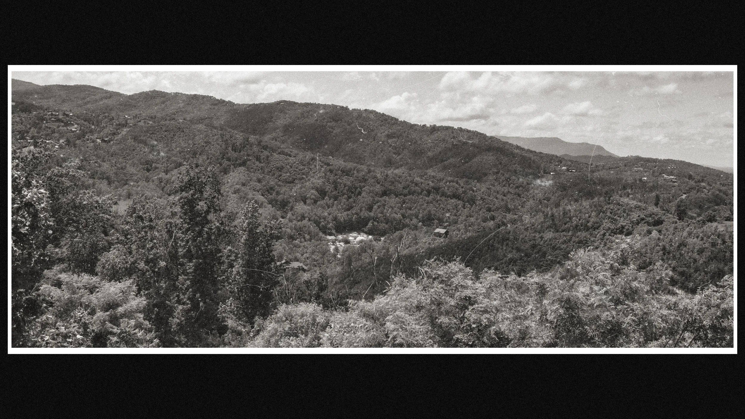 Black and white photo of a mountain landscape with trees, a valley, and scattered houses.
