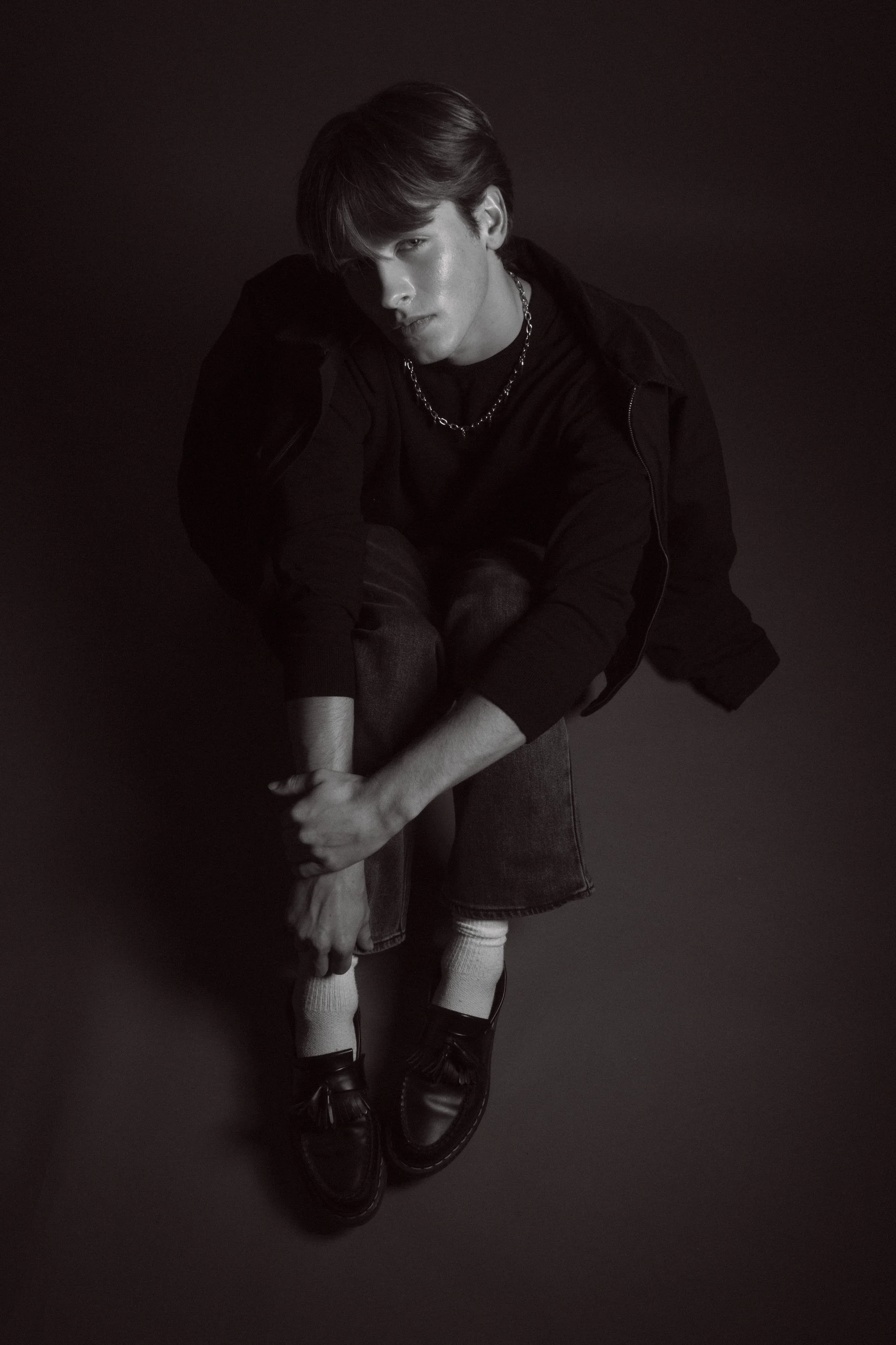 A black-and-white photo of a young man with a haircovering hairstyle, wearing a black shirt, denim shorts, white socks, and black shoes, sitting against a plain backdrop, with a chain necklace visible.